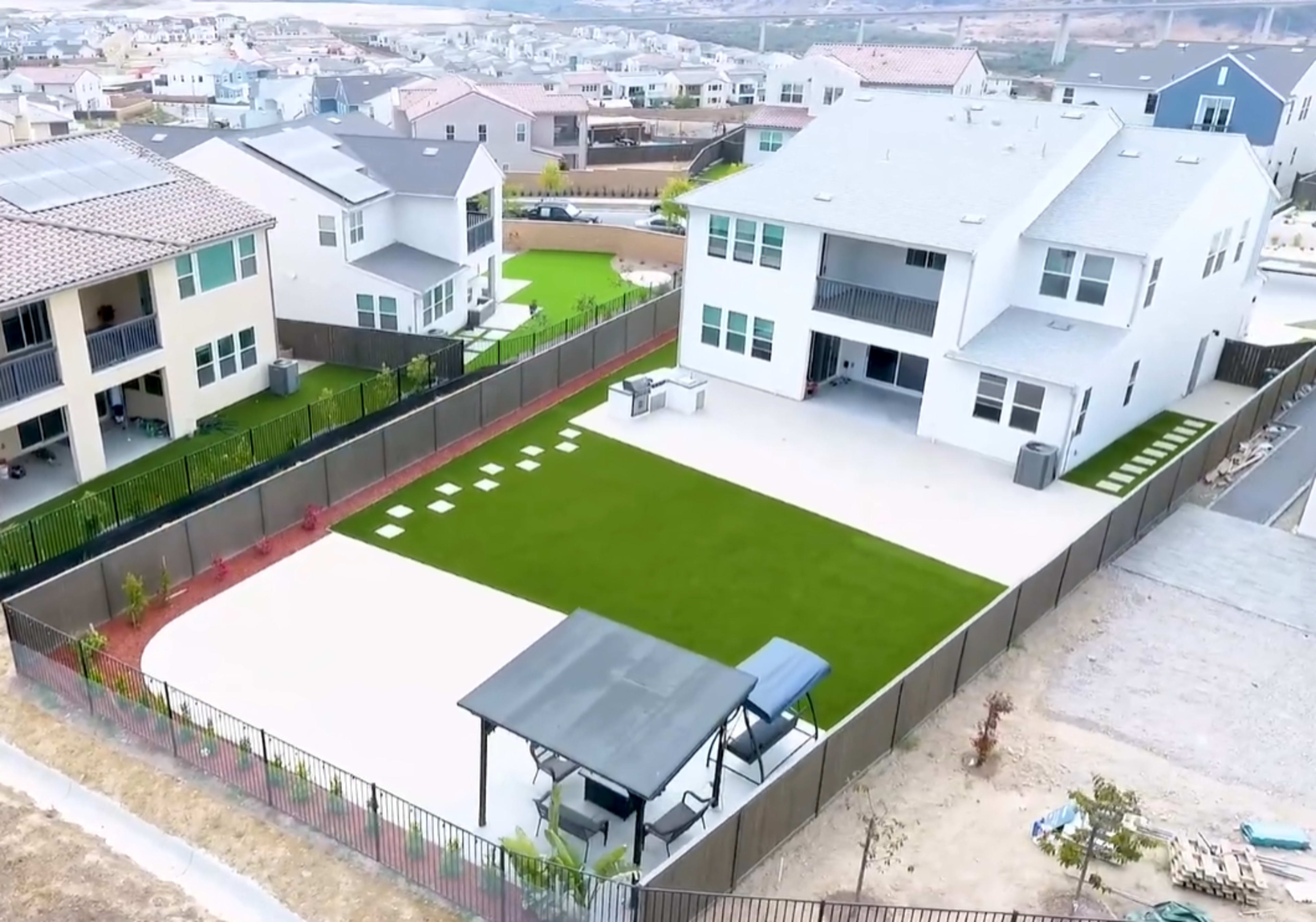 The image shows a large residential backyard featuring artificial grass, a patio area, and a shaded seating structure, surrounded by a fence with views of nearby houses and hills in the background.