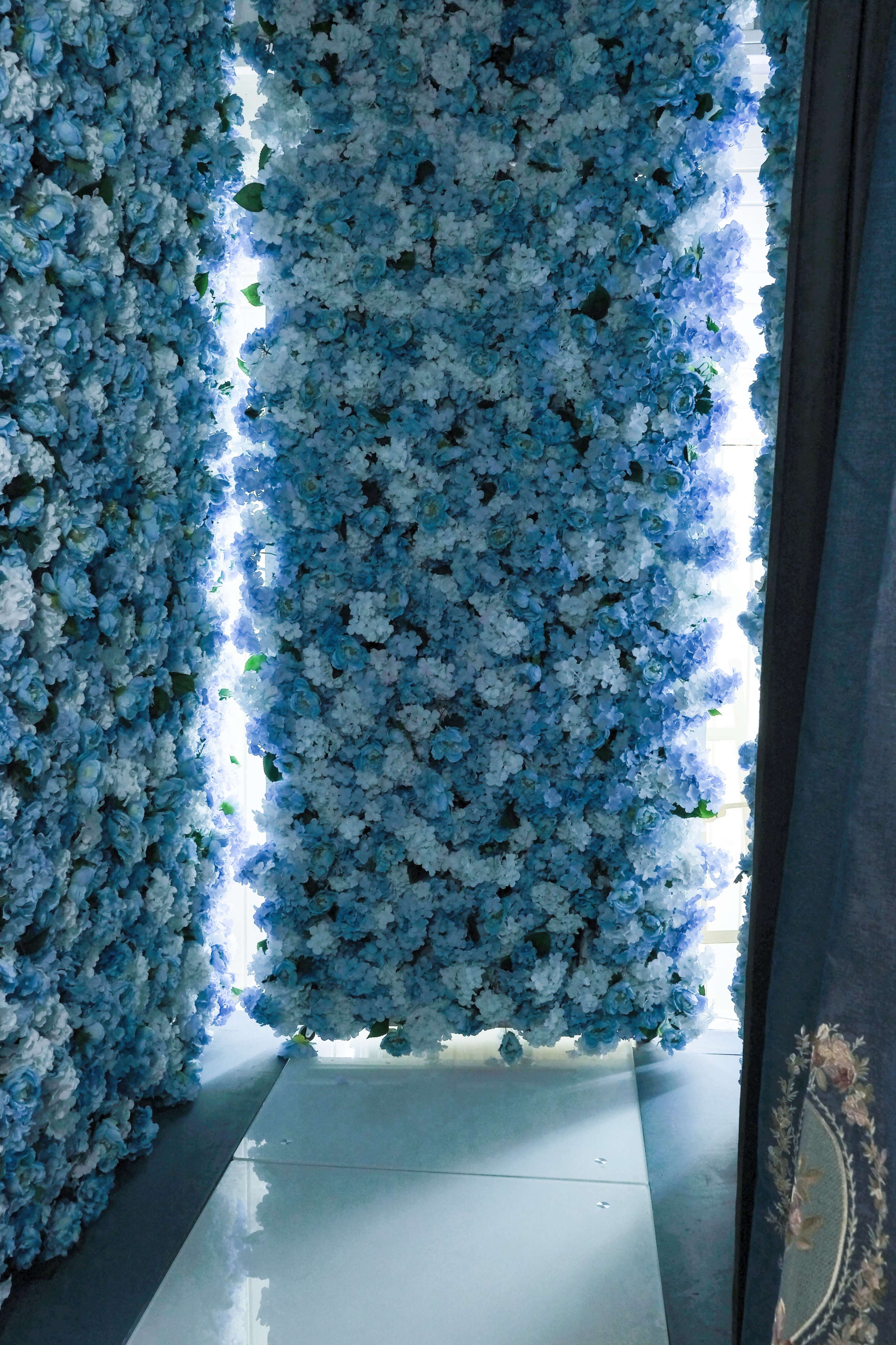 A narrow pathway is flanked by tall walls covered in blue artificial flowers, with bright light illuminating the area.
