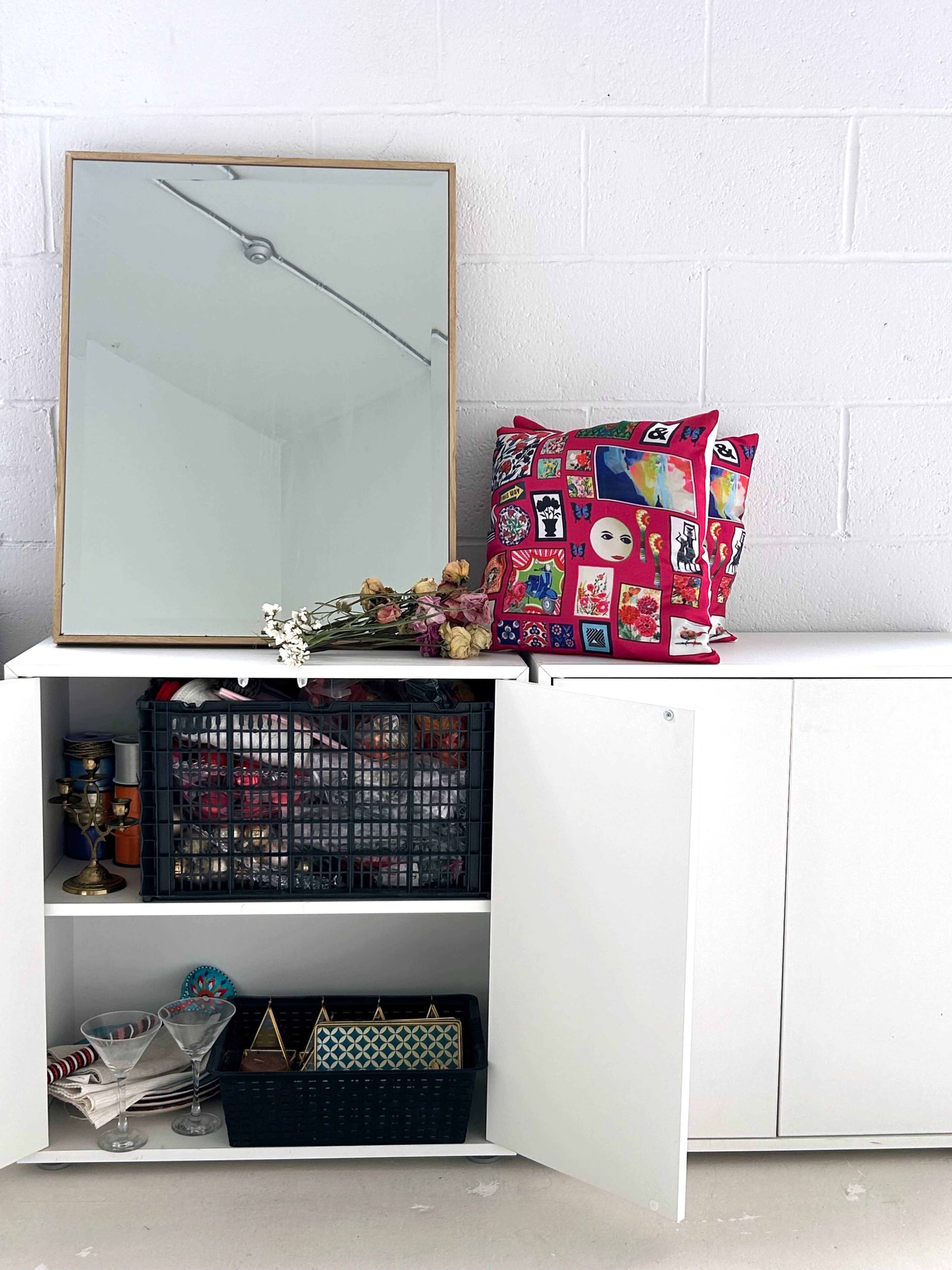 A white cabinet with open doors displays various items, including a black basket and a collection of glassware, while a mirror and a decorative bag sit above it.