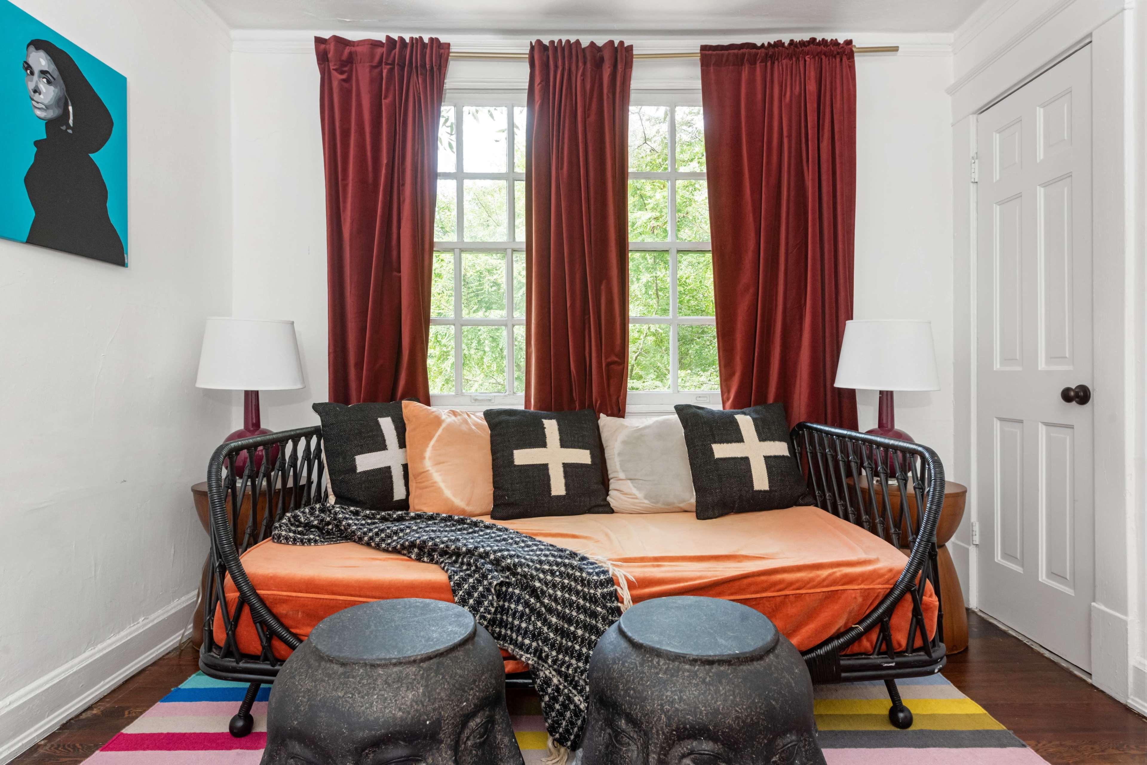 A living room features a daybed with orange bedding, two cross-patterned pillows, black stools, and large windows framed by red curtains.