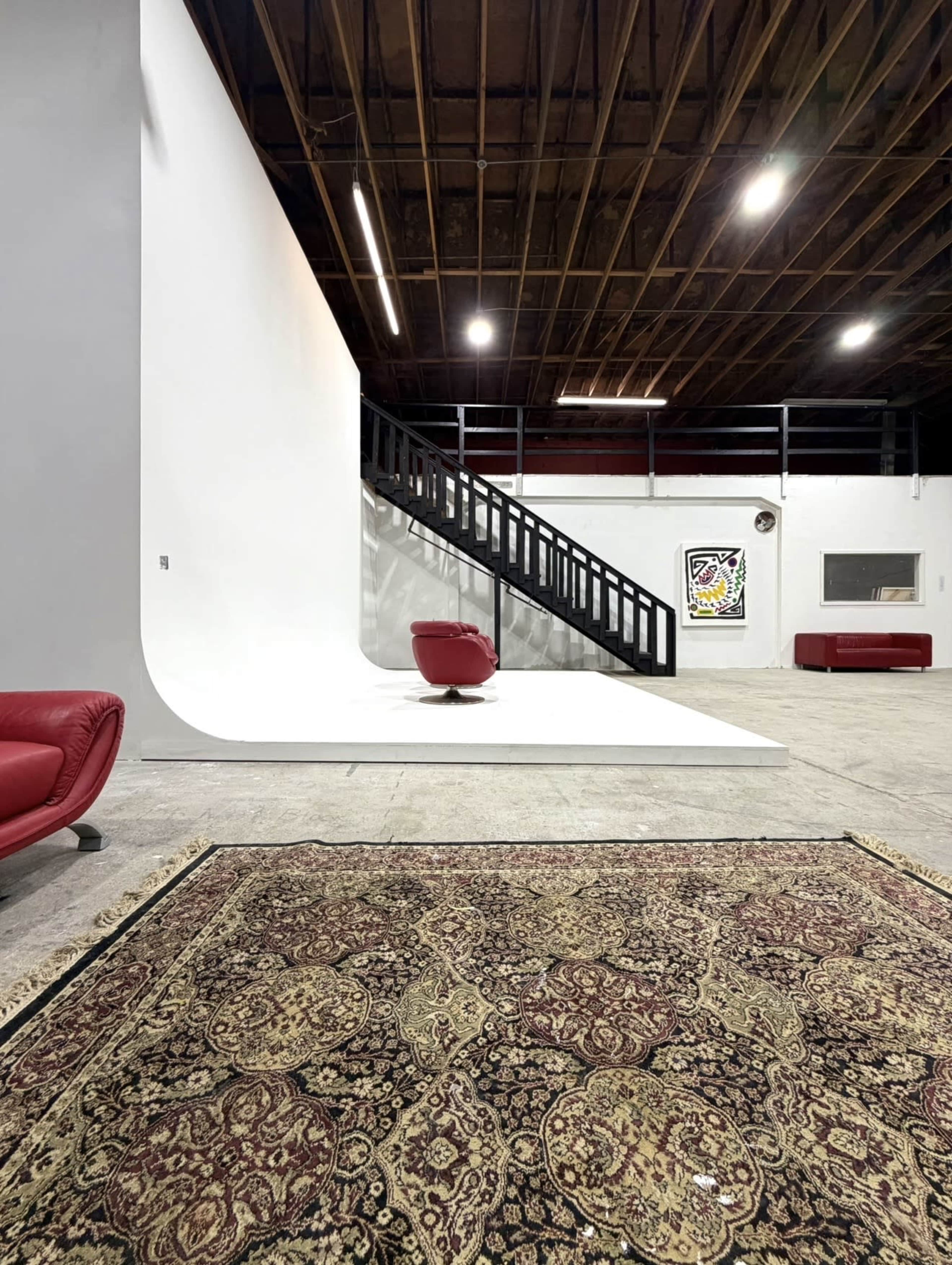 The image shows a spacious interior with a red chair on a curved white backdrop, a staircase against the wall, and a patterned rug on the floor.