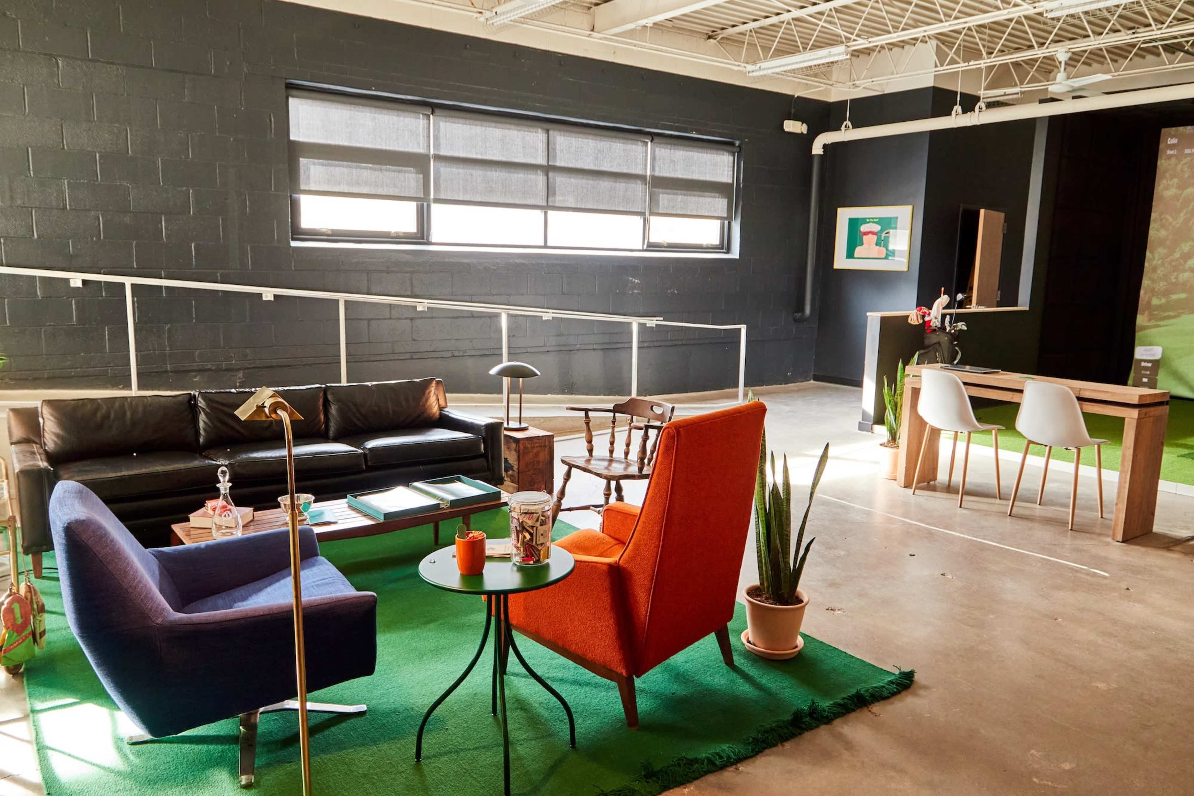 The image shows a modern interior featuring a black couch, an orange chair, a blue chair, and a small round table on green carpet, with large windows and a minimalist decor.