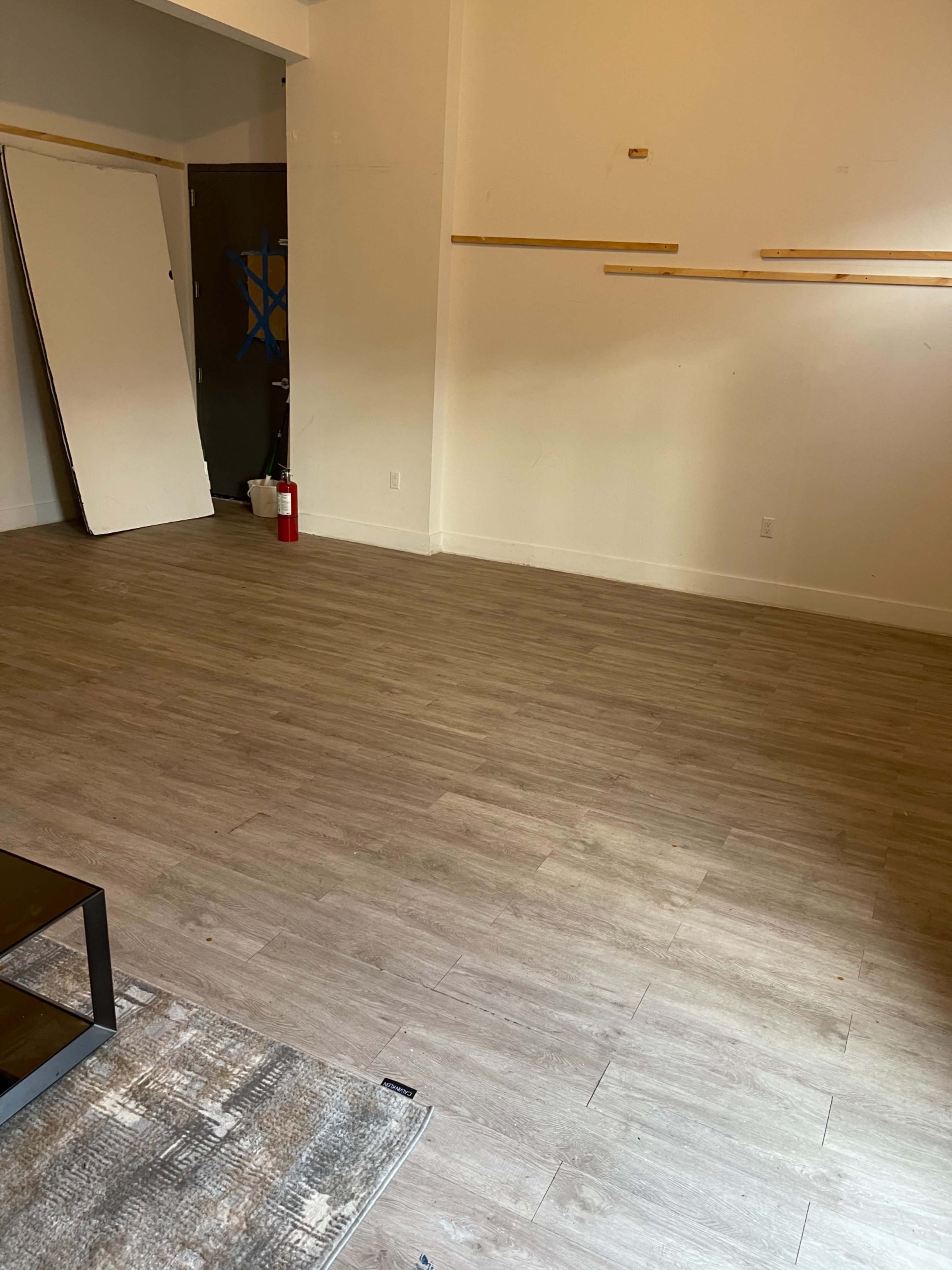 The image shows a spacious, empty room with light wooden flooring and a large blank wall, featuring a neatly arranged fire extinguisher in the corner.