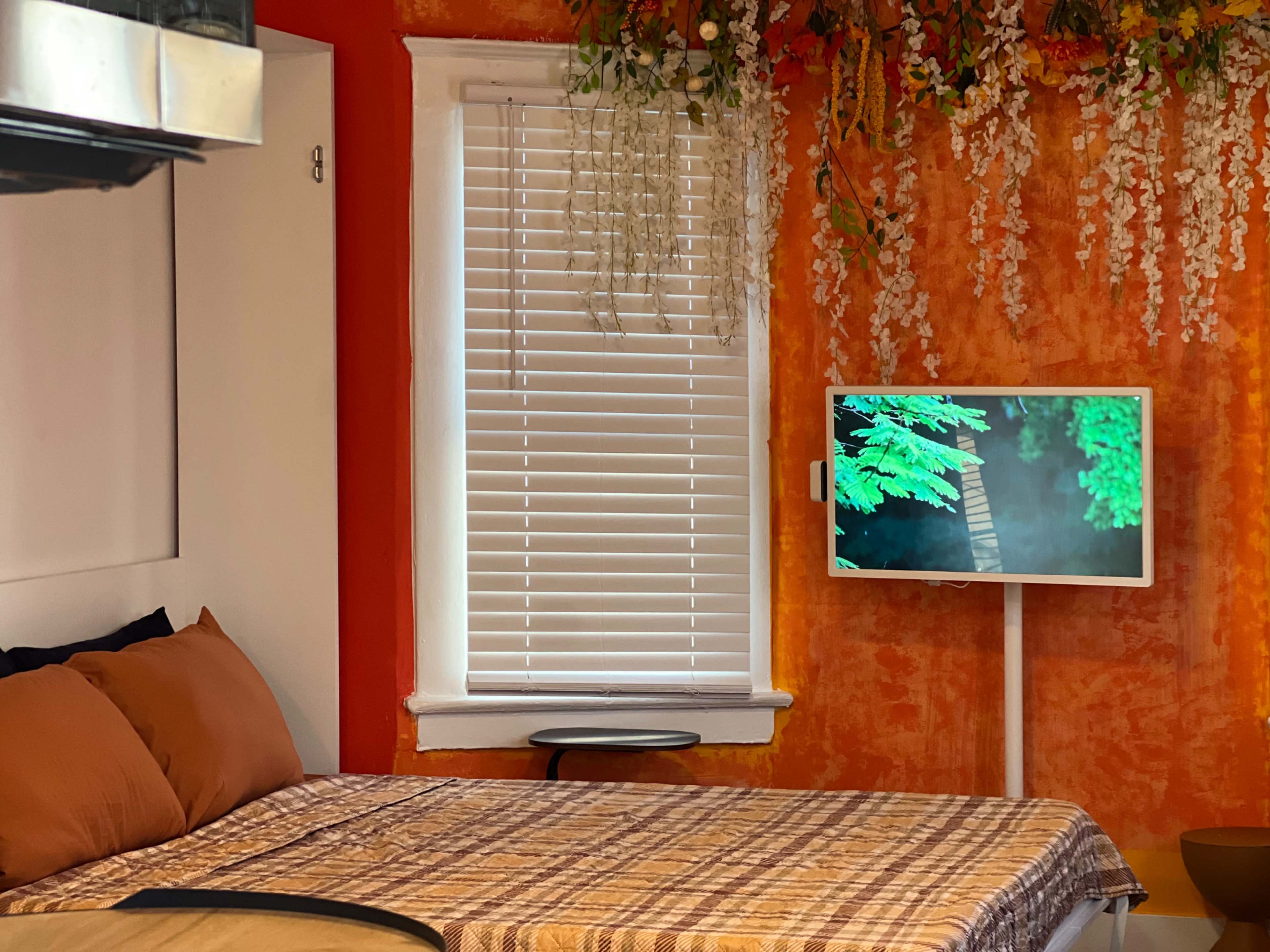 The image shows a bedroom with a bed featuring brown pillows, a modern TV mounted on the wall displaying a nature scene, and an orange wall adorned with decorative plants.