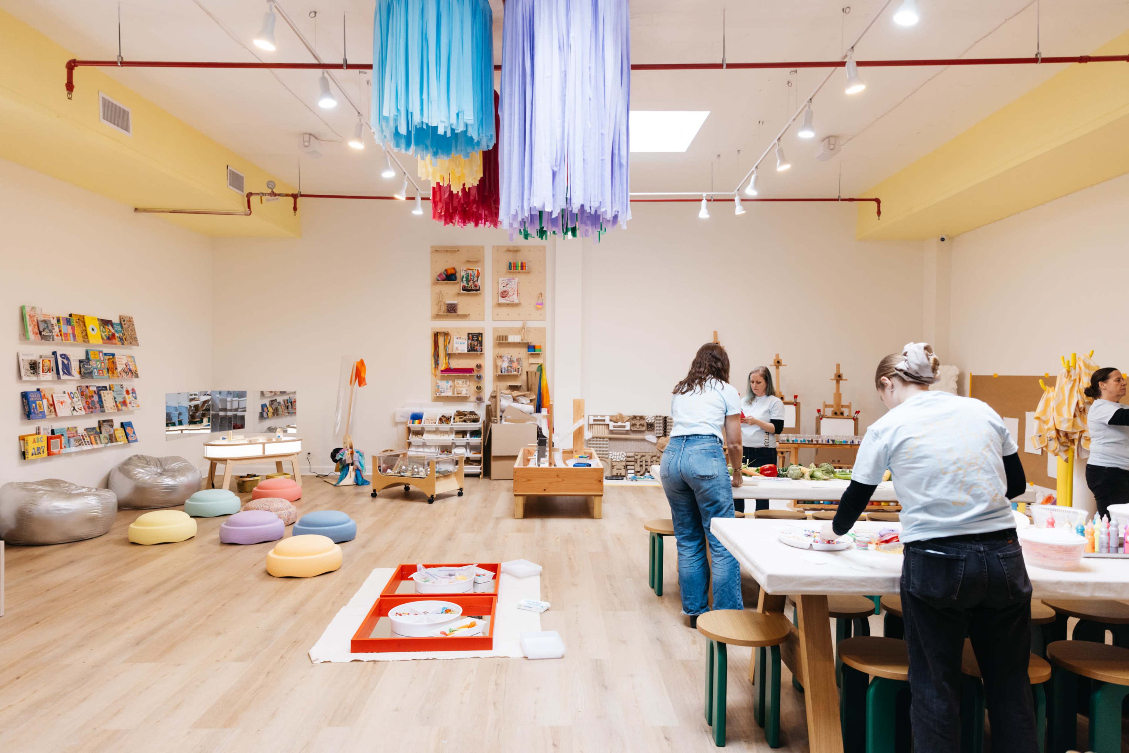 The image shows a brightly lit art studio with colorful decorations, tables set up for creative activities, and various craft supplies arranged around the space.