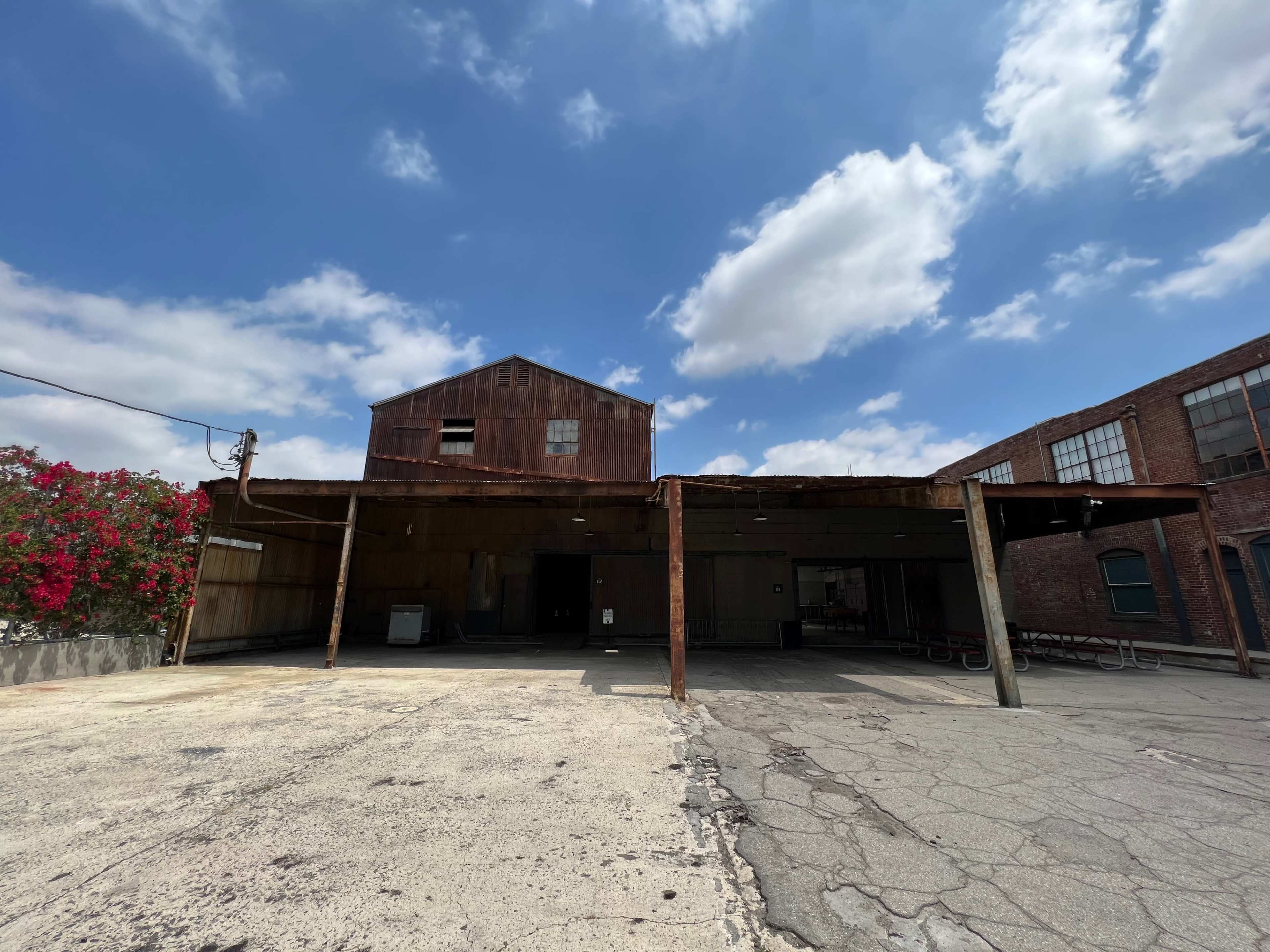 An abandoned warehouse with a rusted metal structure and a concrete lot beneath a partly cloudy sky.