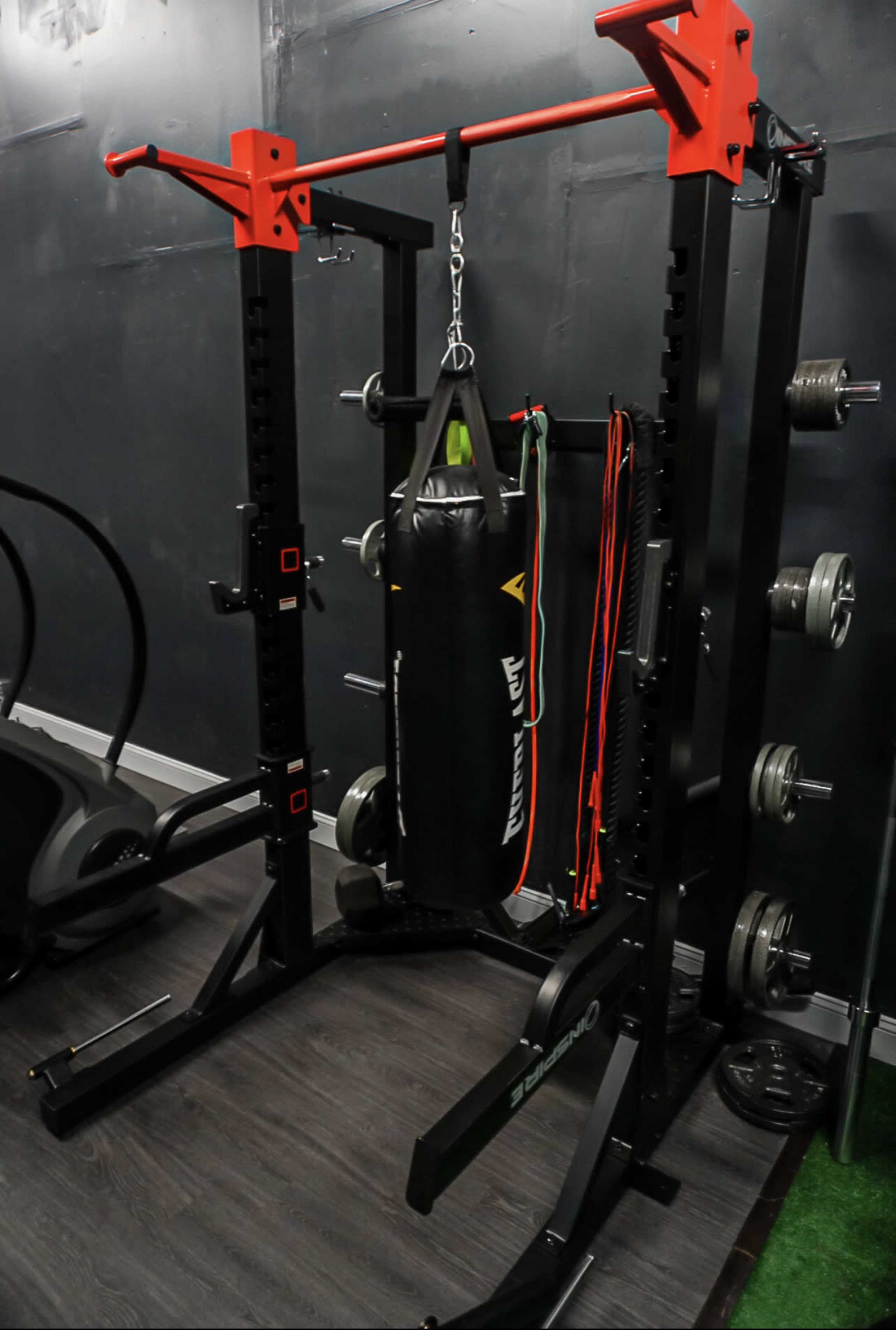 A black and red power rack with a hanging punching bag, resistance bands, and weight plates, set against a dark wall in a gym.