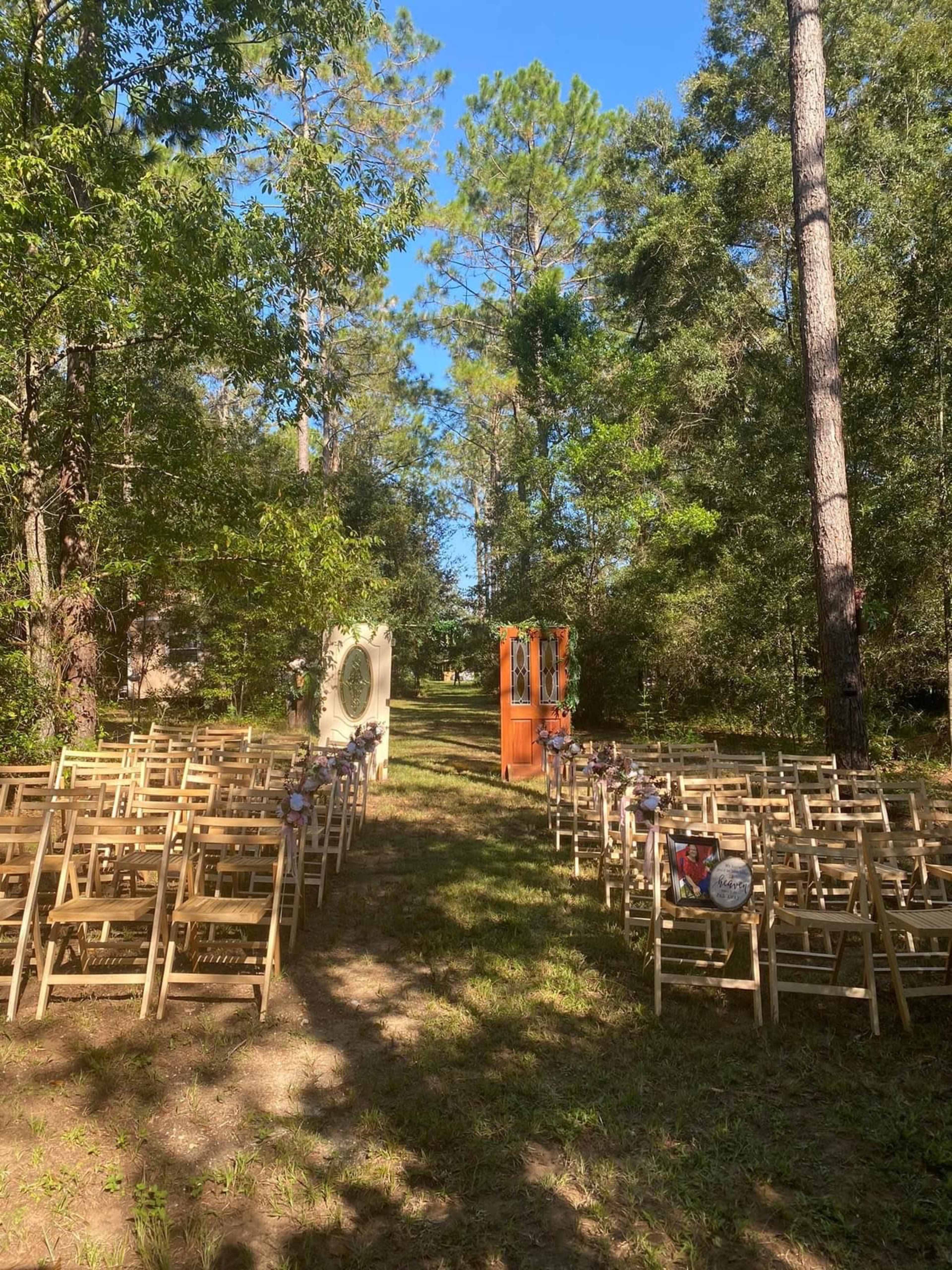 The image shows an outdoor wedding ceremony setup with rows of wooden chairs facing a decorative arch and an orange door, surrounded by trees.