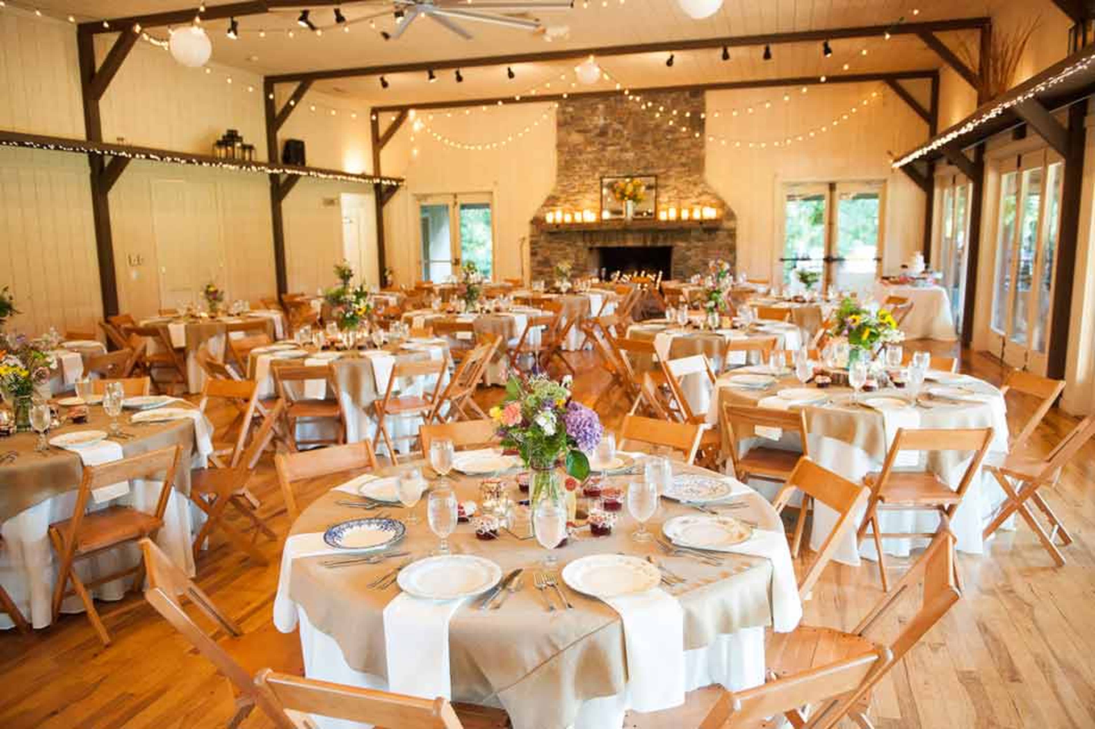 A spacious event venue features several elegantly arranged tables with floral centerpieces, surrounded by wooden chairs and a stone fireplace.
