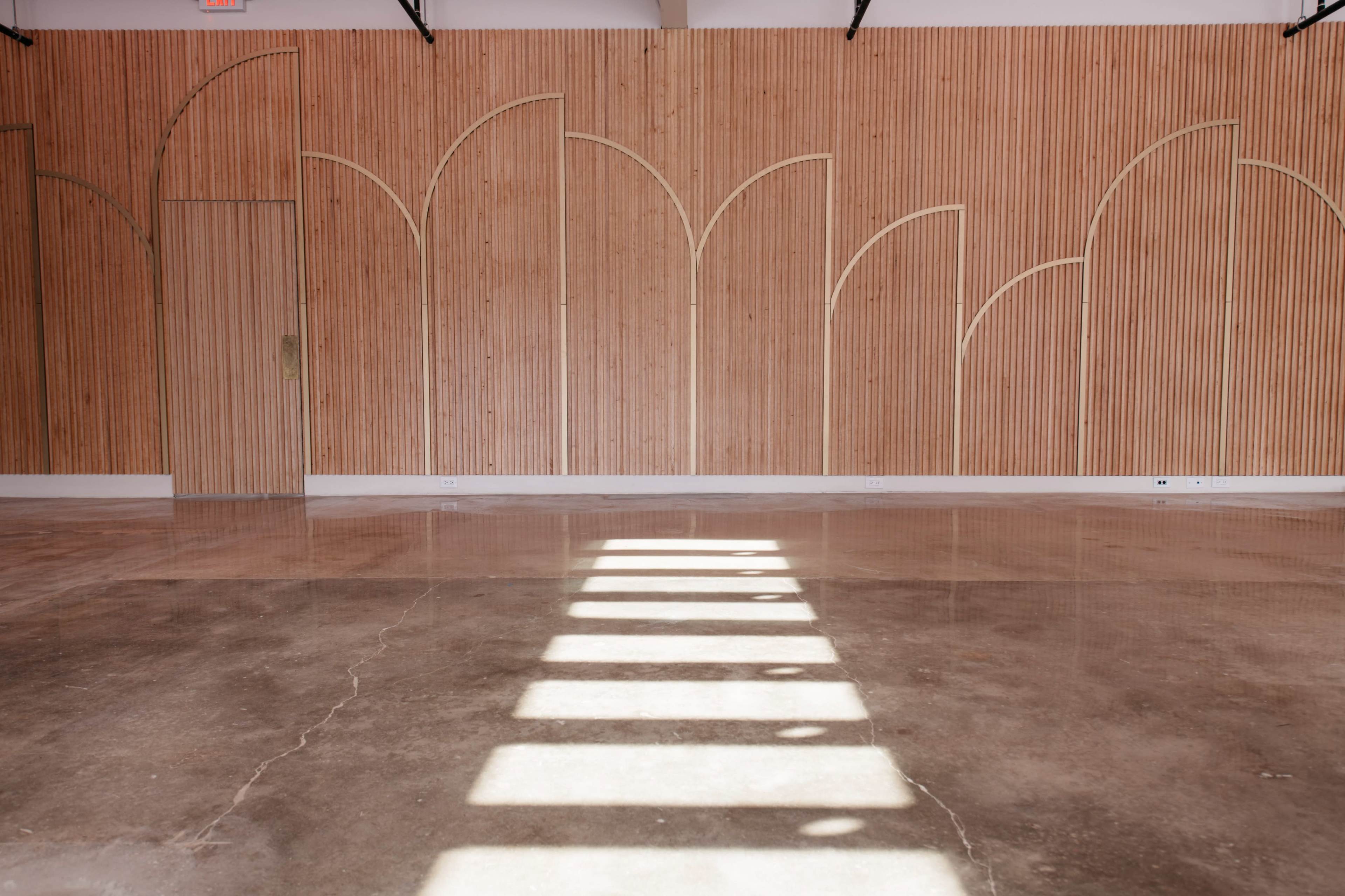 The image shows a spacious interior with a wooden paneled wall featuring an arched design and concrete flooring reflecting strong sunlight.