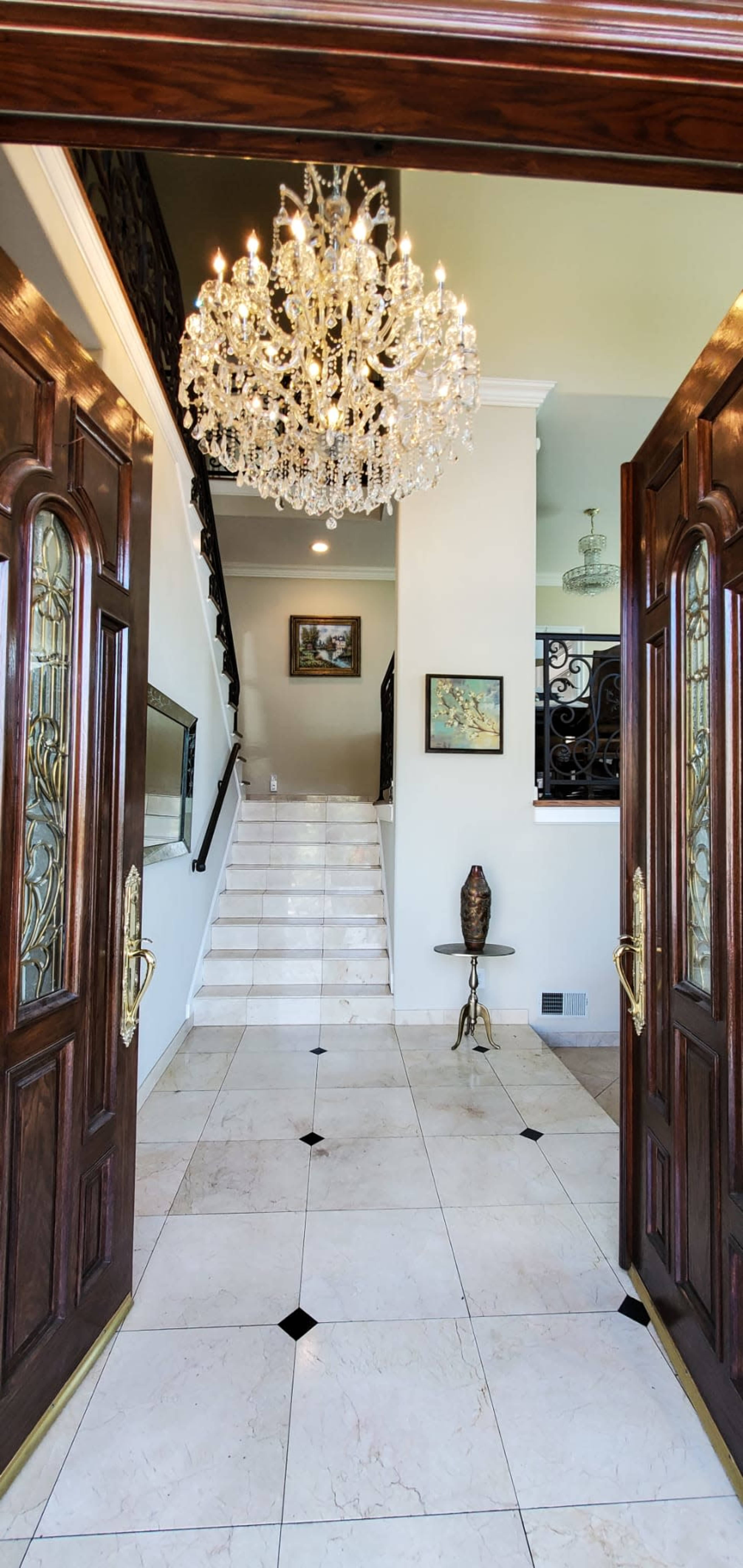 A spacious foyer with double wooden doors, a chandelier hanging from the ceiling, and a staircase leading to an upper level, accompanied by decorative artwork and a small side table.