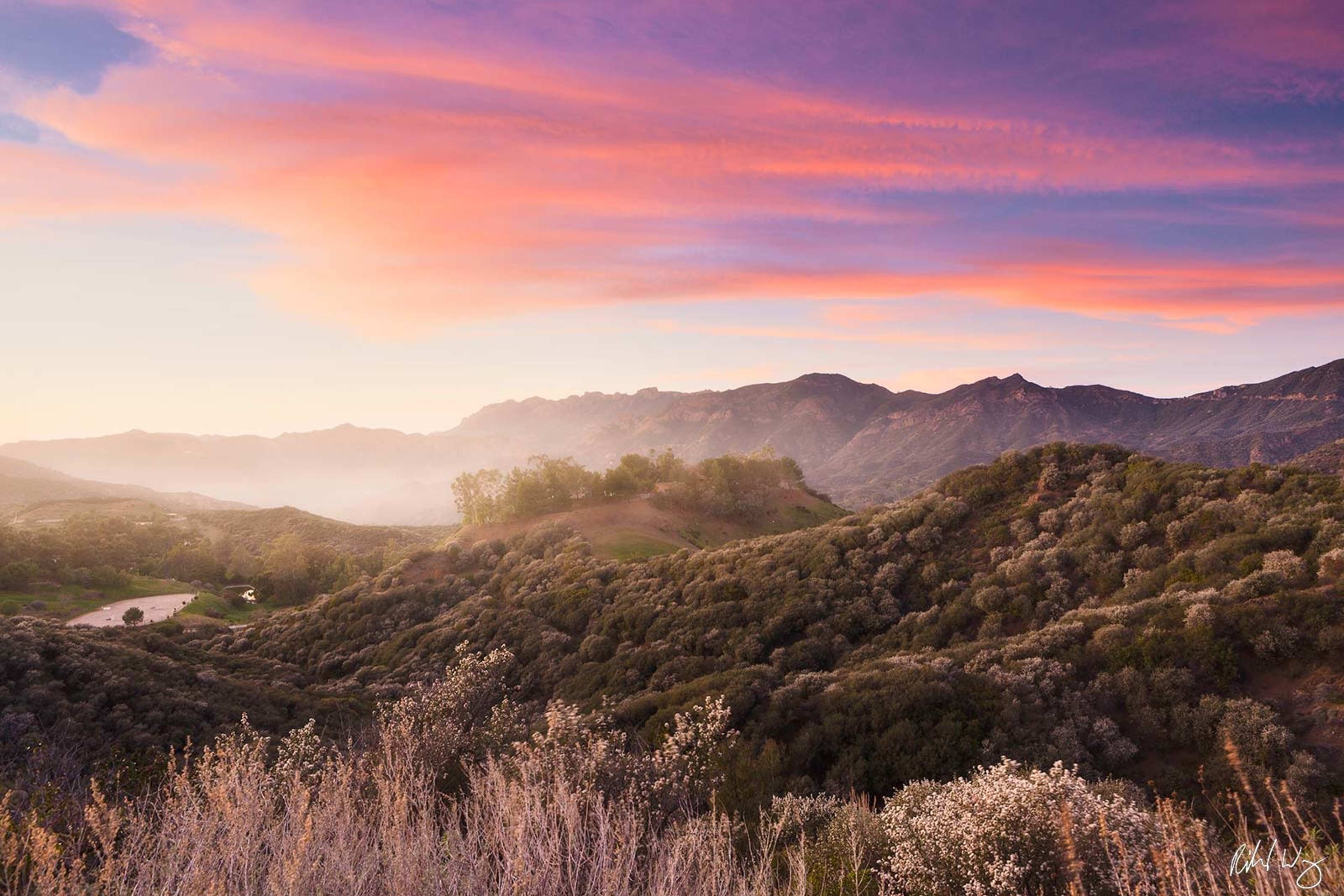 The image shows a scenic view of rolling hills under a colorful sky at sunset, with a faint mist in the distance.