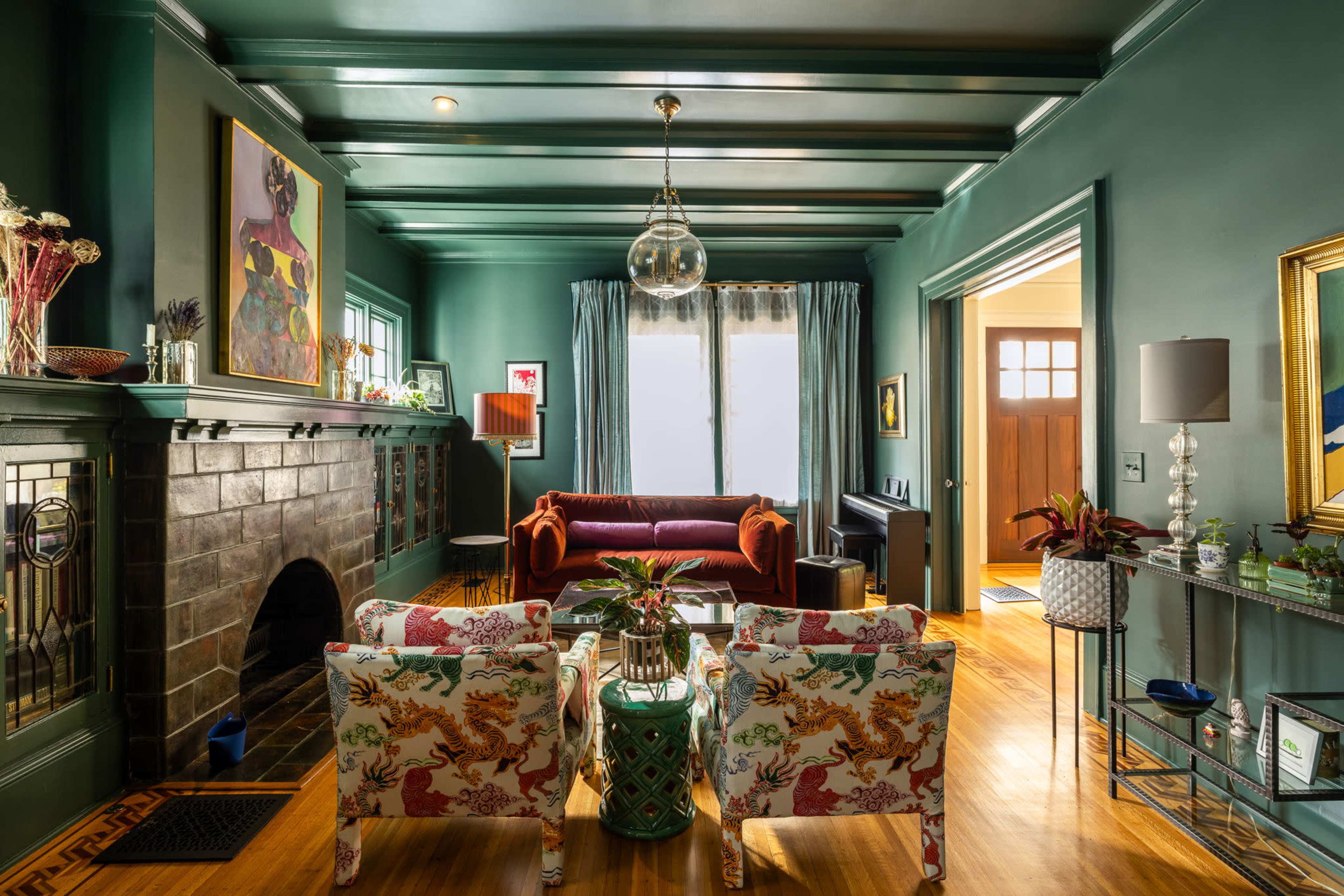 The image shows a living room featuring a dark green color scheme, a fireplace with decorative tiles, two distinctive armchairs, and a mix of contemporary and classic artwork.