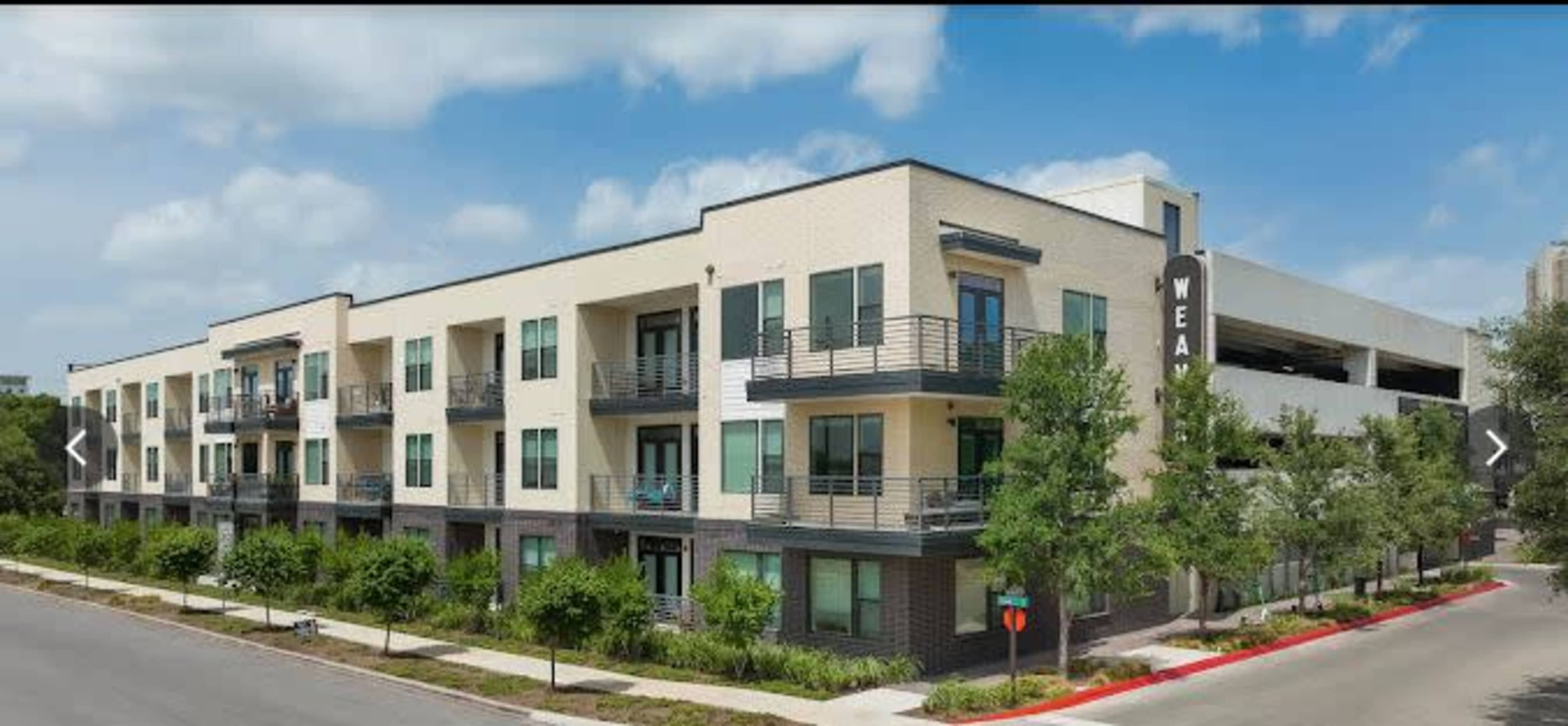 A modern, three-story apartment building features multiple balconies and large windows, surrounded by landscaped greenery.
