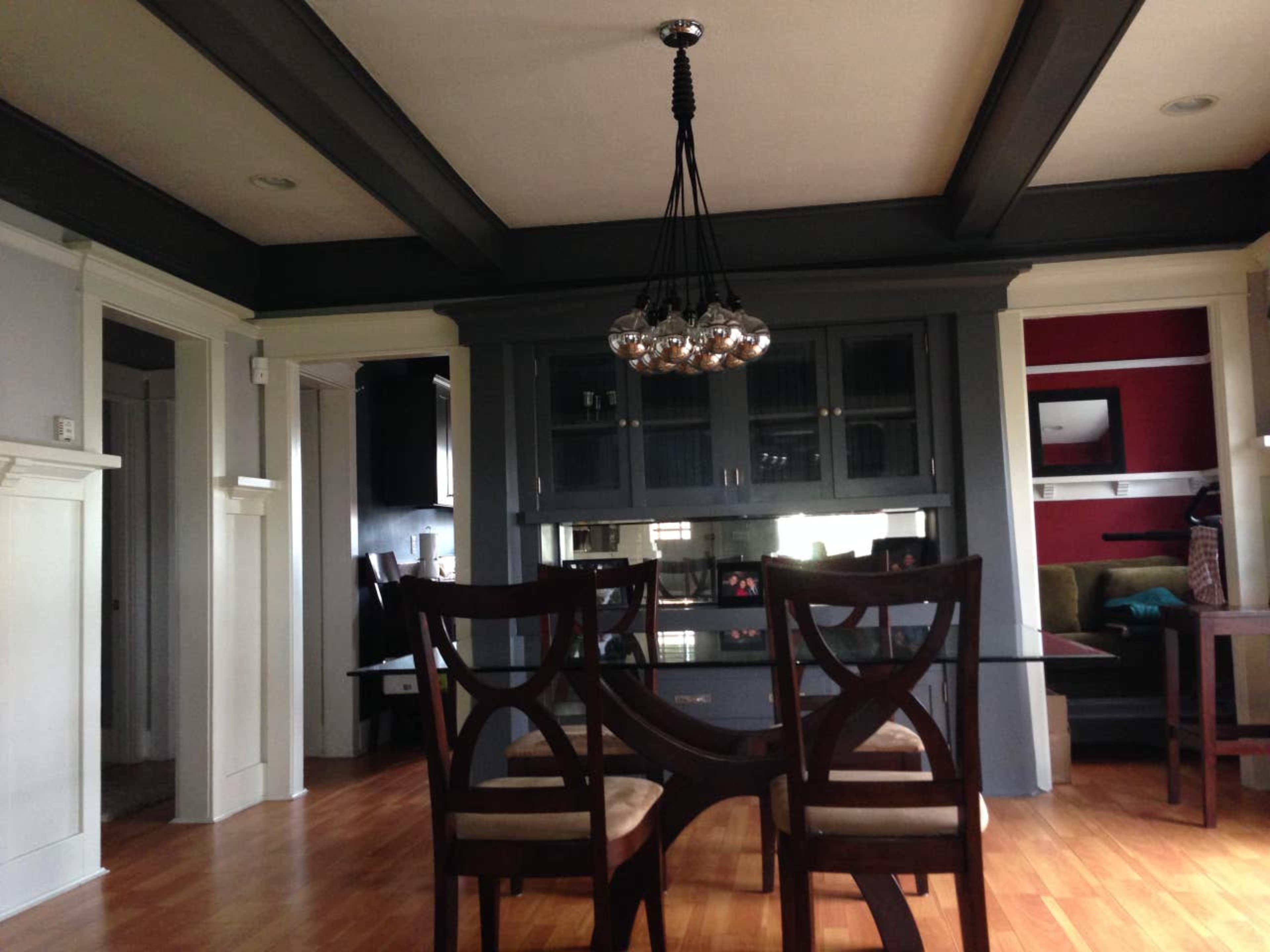 A dining area with a wooden table and chairs, featuring a chandelier and a wall unit, adjacent to a kitchen and a living space.