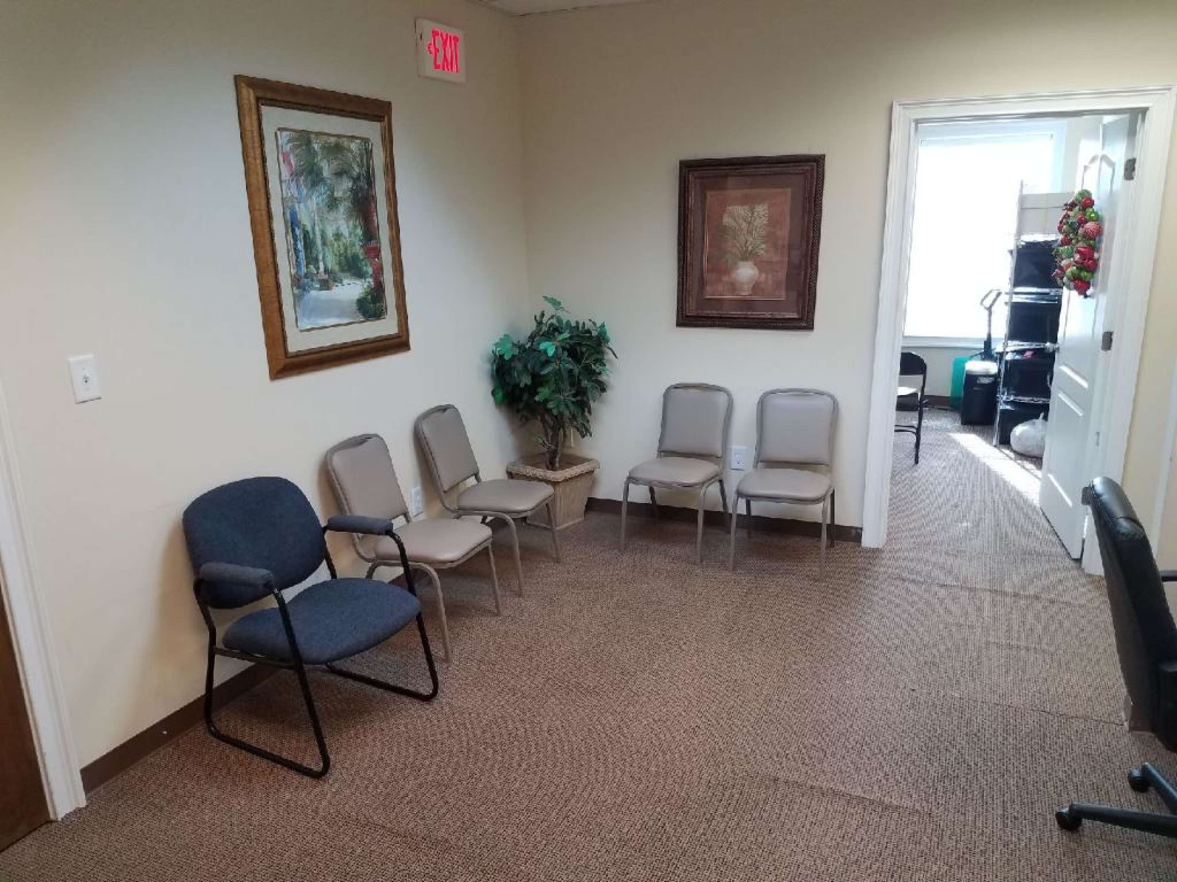 A waiting room with two chairs against one wall, a small plant, and a framed painting, leading to a door and a glimpse of another room.