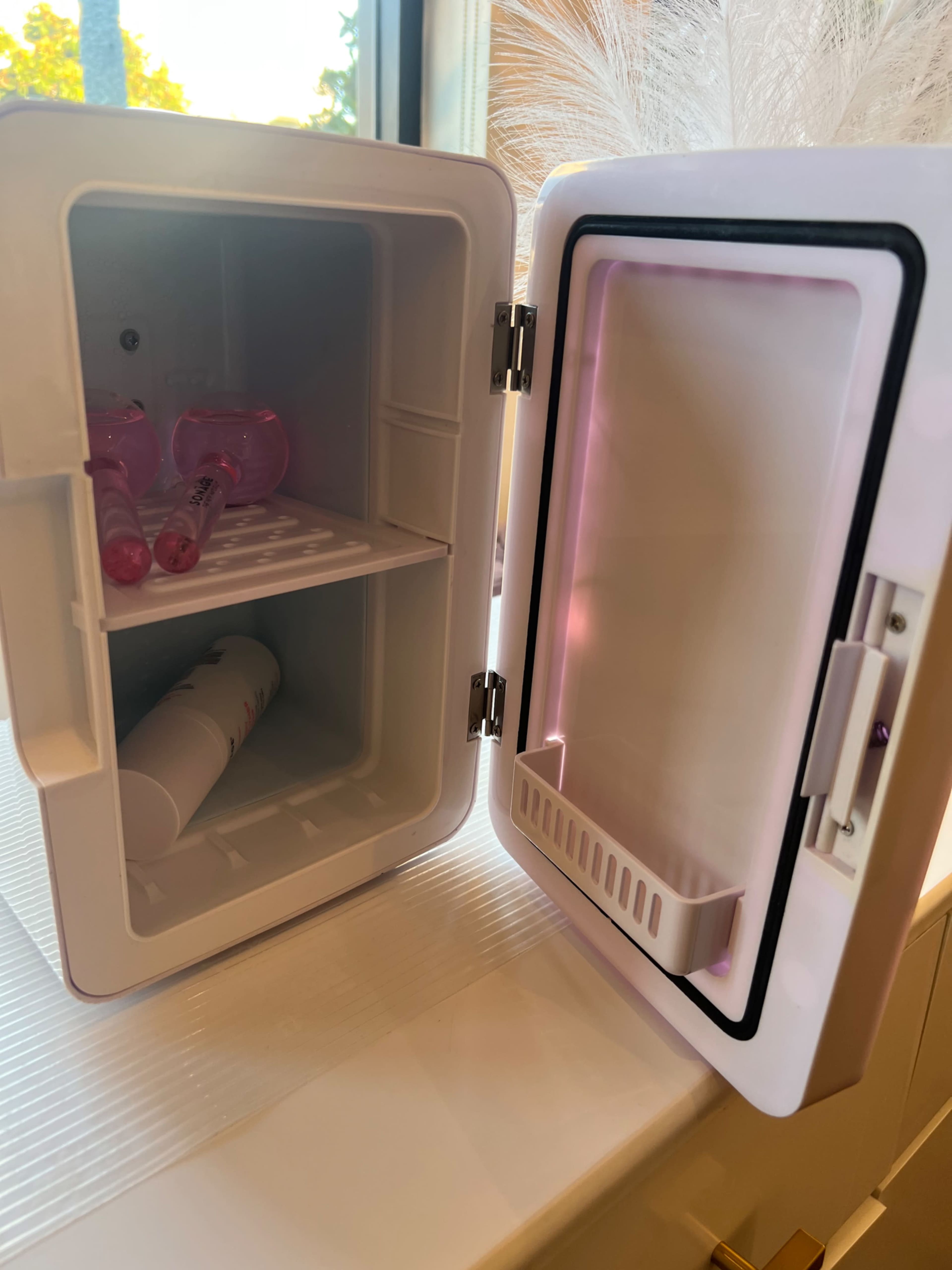 The image shows an open small refrigerator with a white interior, featuring two shelves and some cosmetic containers inside.