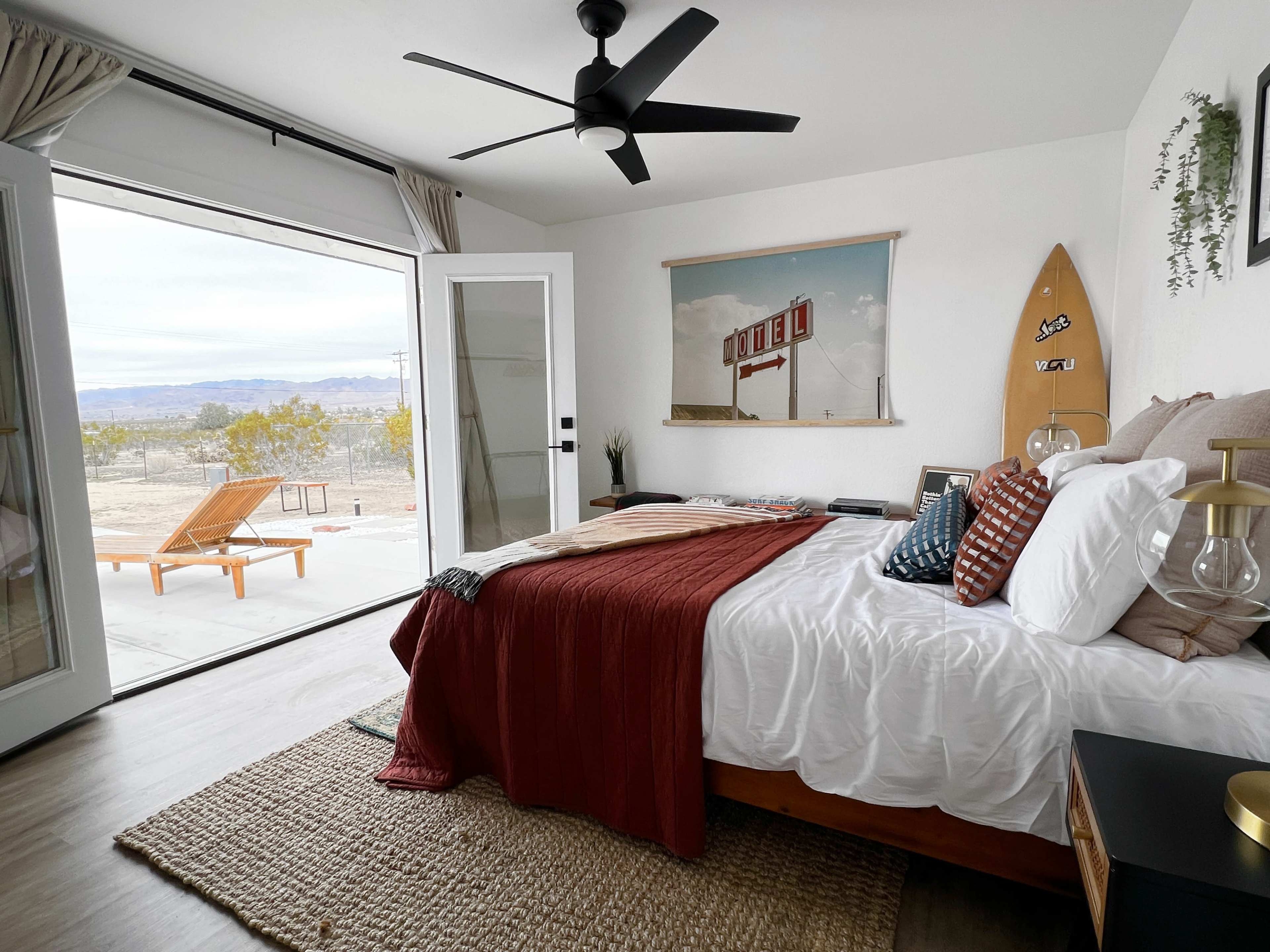 A cozy bedroom features a large window opening to a desert view, with a surfboard, bed, and modern furnishings.