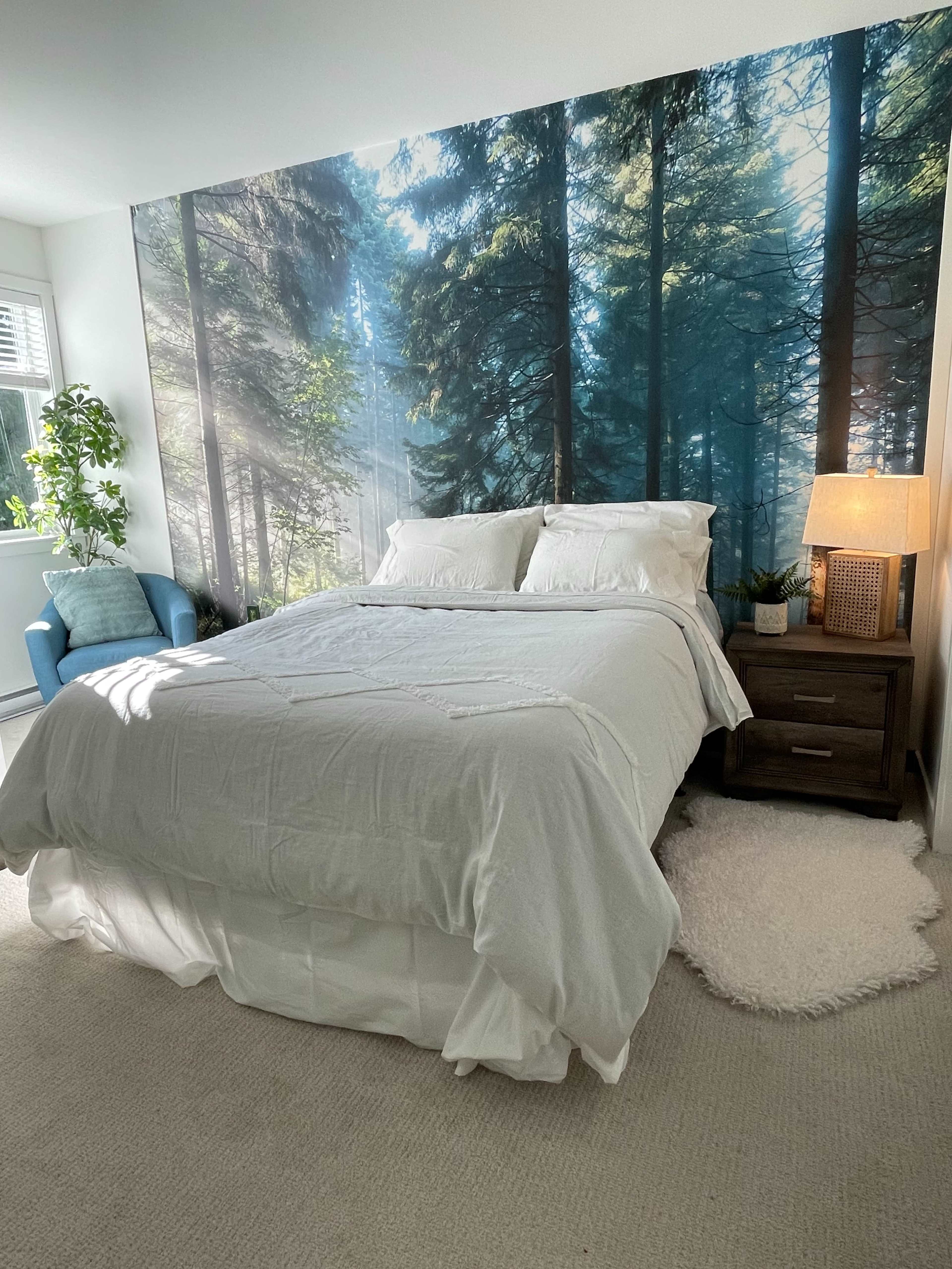A bedroom features a bed with white bedding, a nightstand with a lamp, a blue chair, and a large forest mural on the wall.