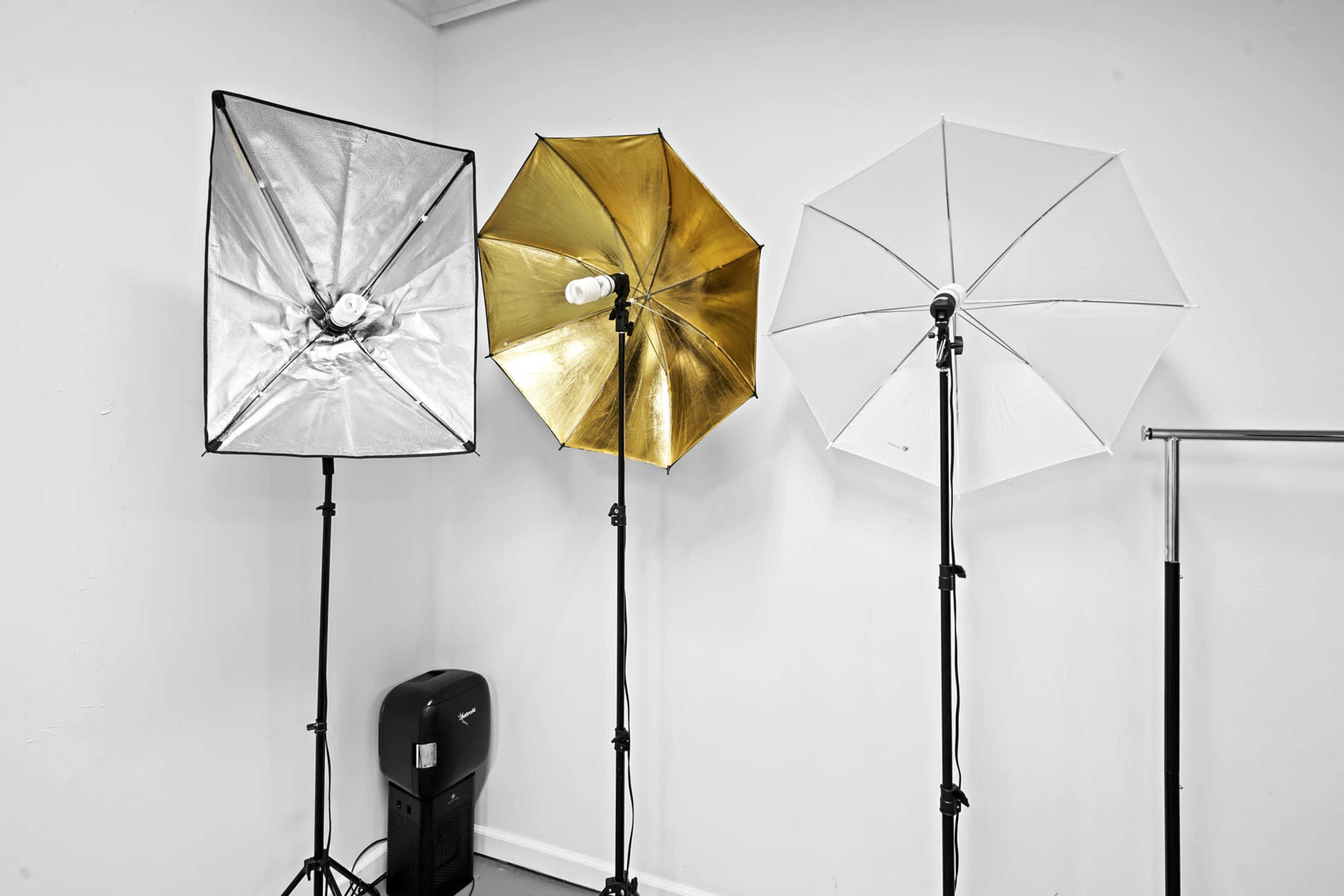 Three studio light umbrellas on stands, with a black and silver umbrella on the left, a gold umbrella in the center, and a white umbrella on the right, alongside a black equipment cart in the background.
