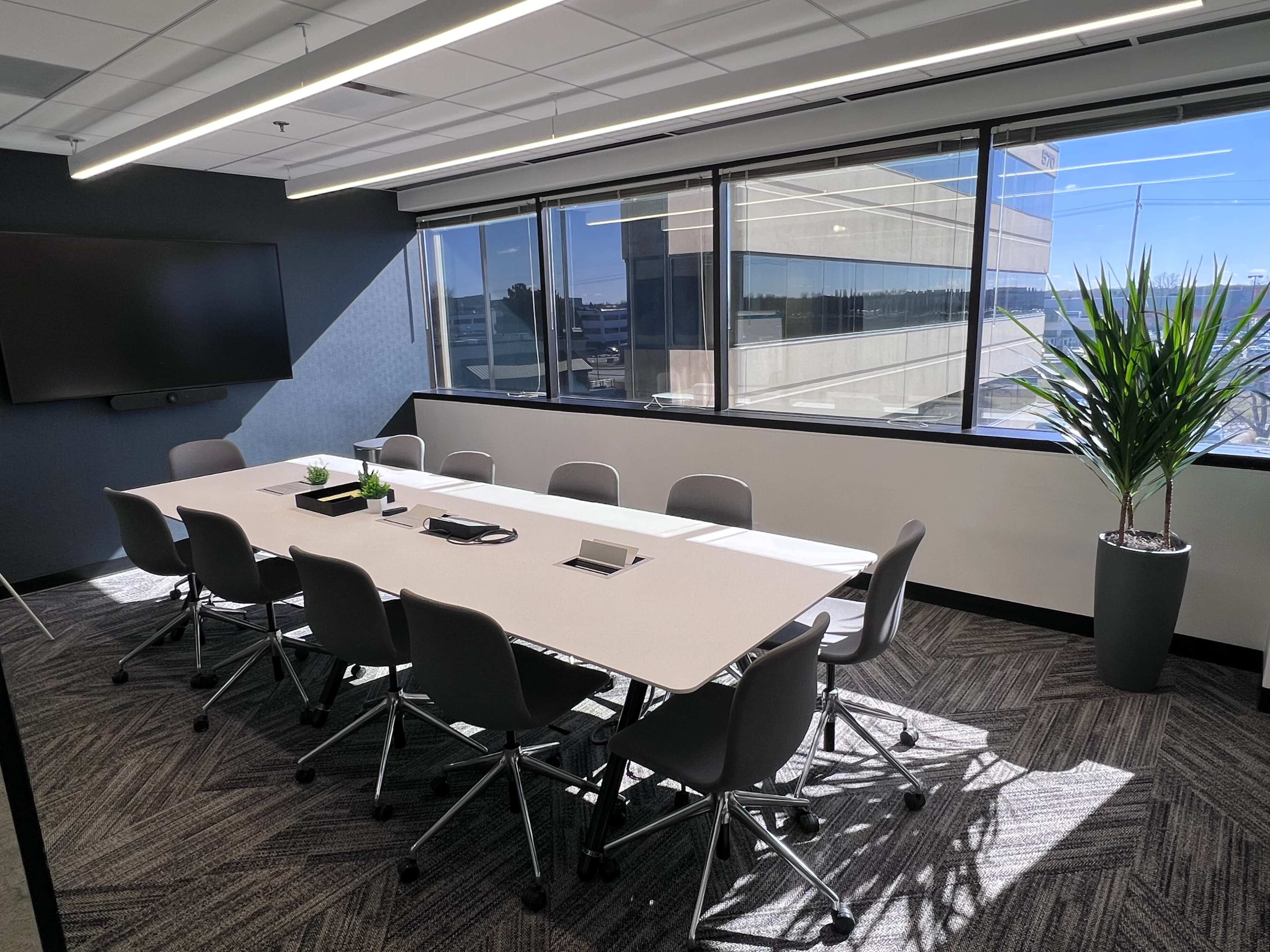 A modern conference room features a long table surrounded by ergonomic chairs, with large windows offering a view of a cityscape.