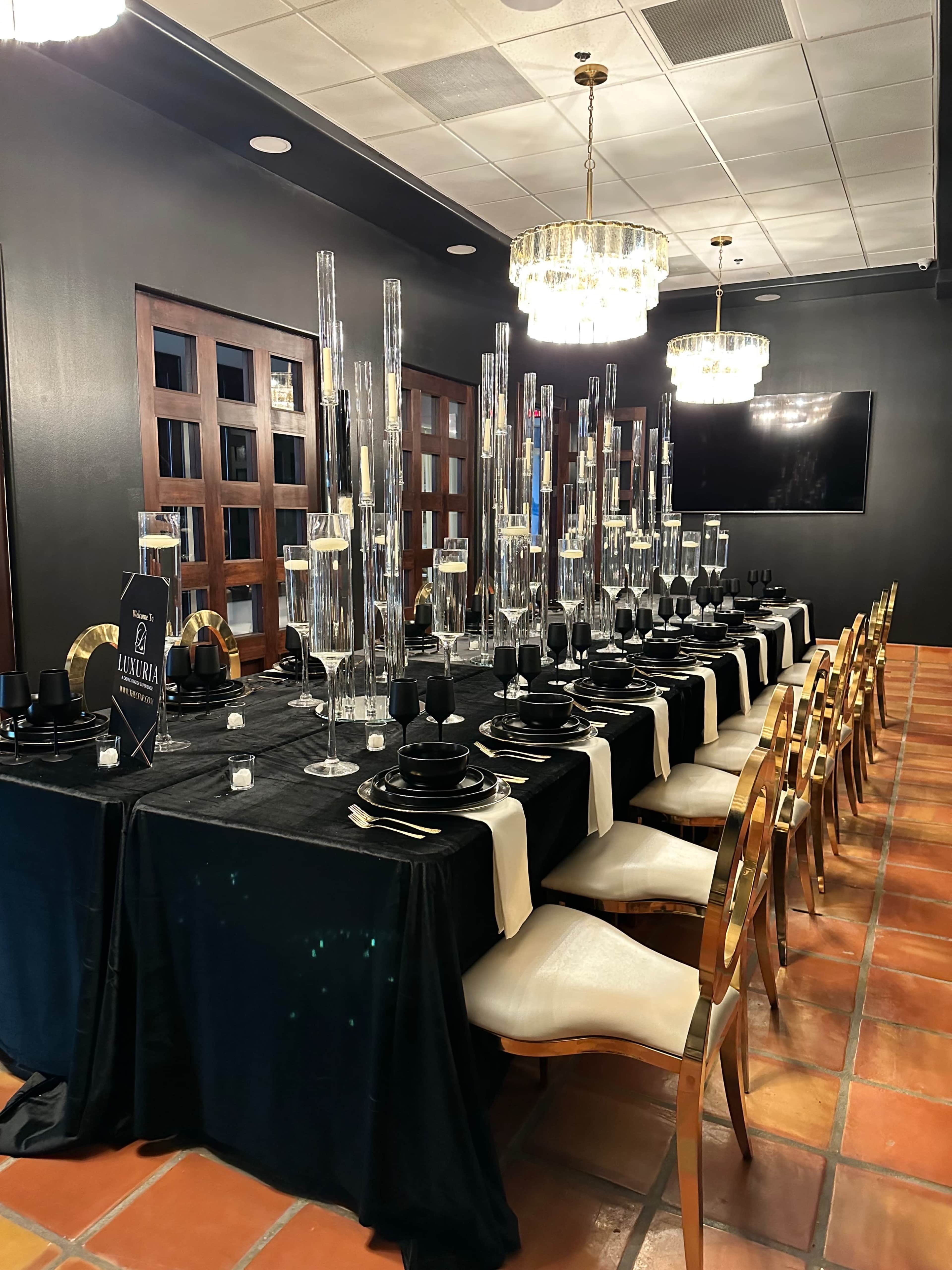 A long, elegantly set dining table with black and gold tableware, surrounded by tall candle holders, in a dimly lit room.