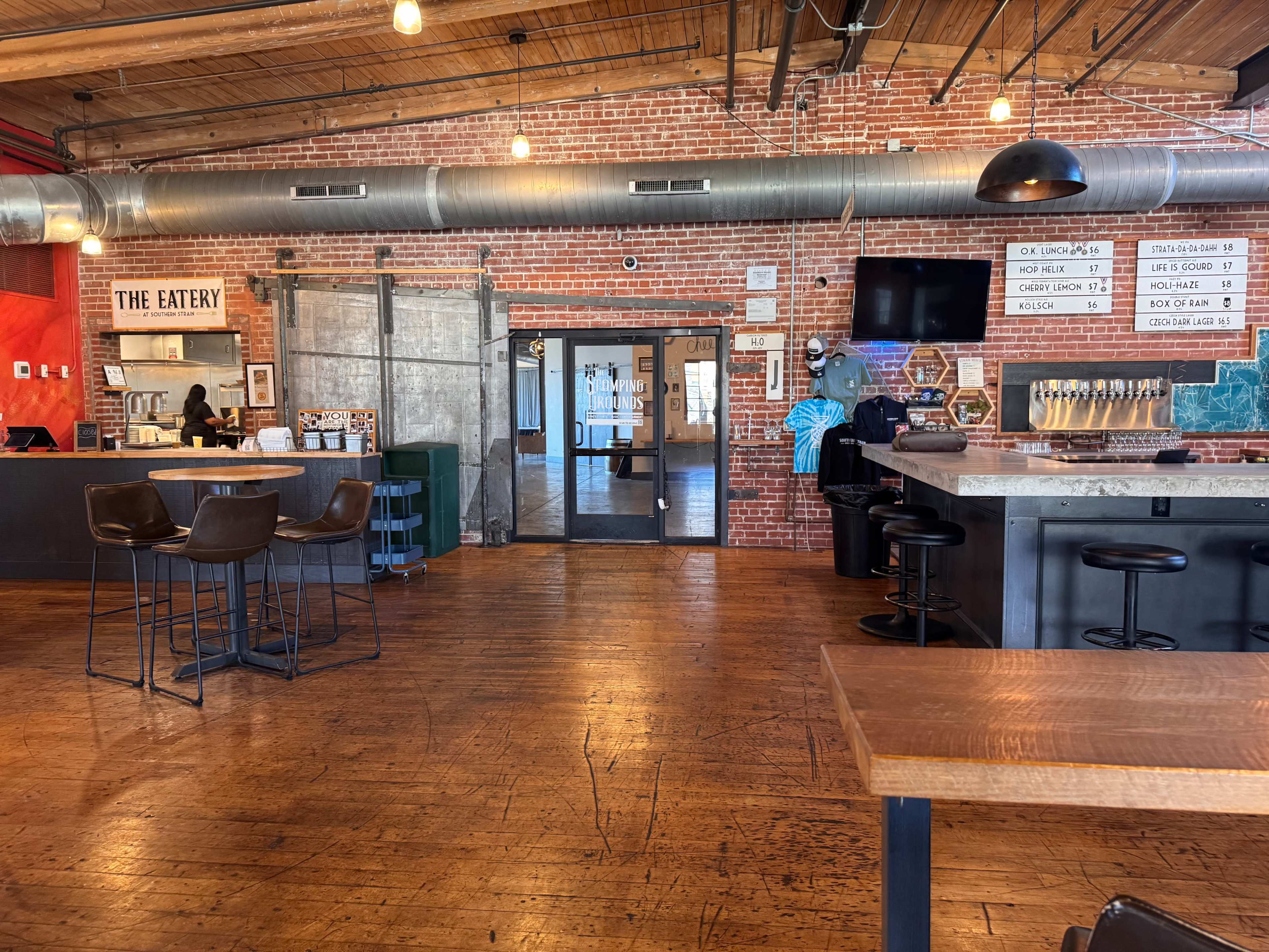 The interior of a cafe features exposed brick walls, wooden floors, and a counter with seating and a large menu displayed on the wall.