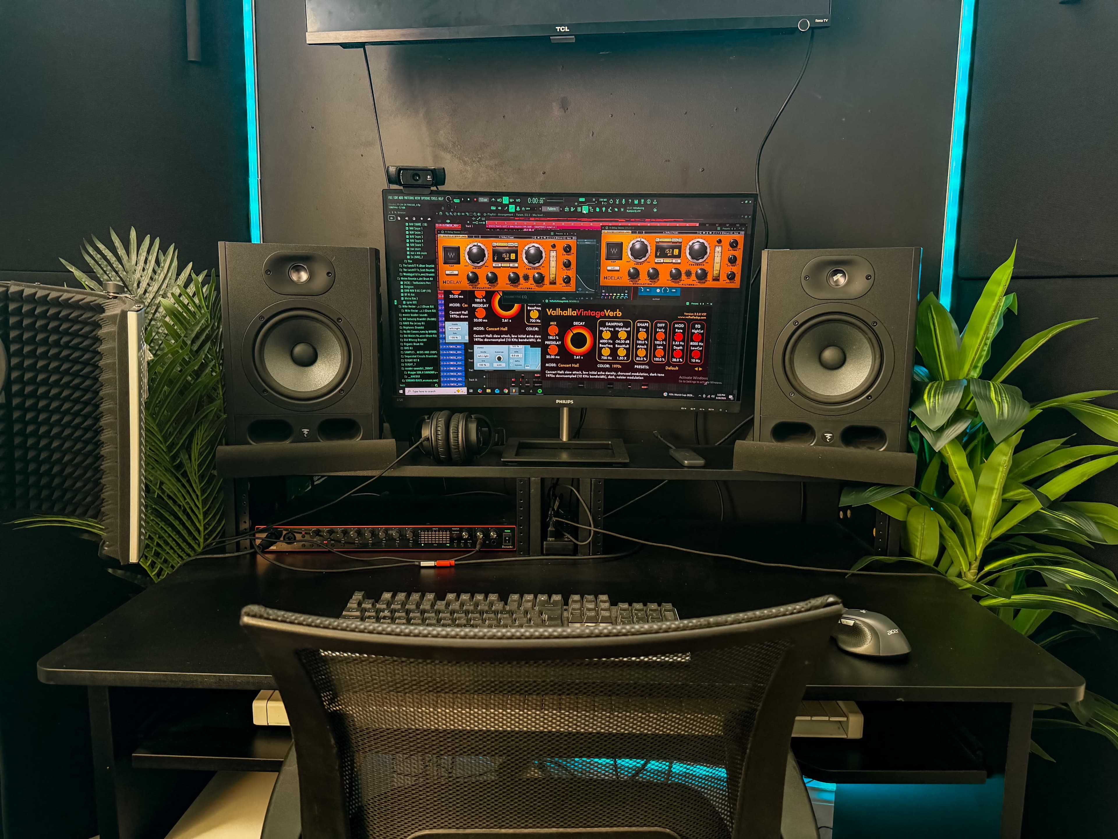 The image shows a music production workspace featuring a computer monitor with audio software displayed, two large speakers on either side, a keyboard, mouse, and some plants in the background.