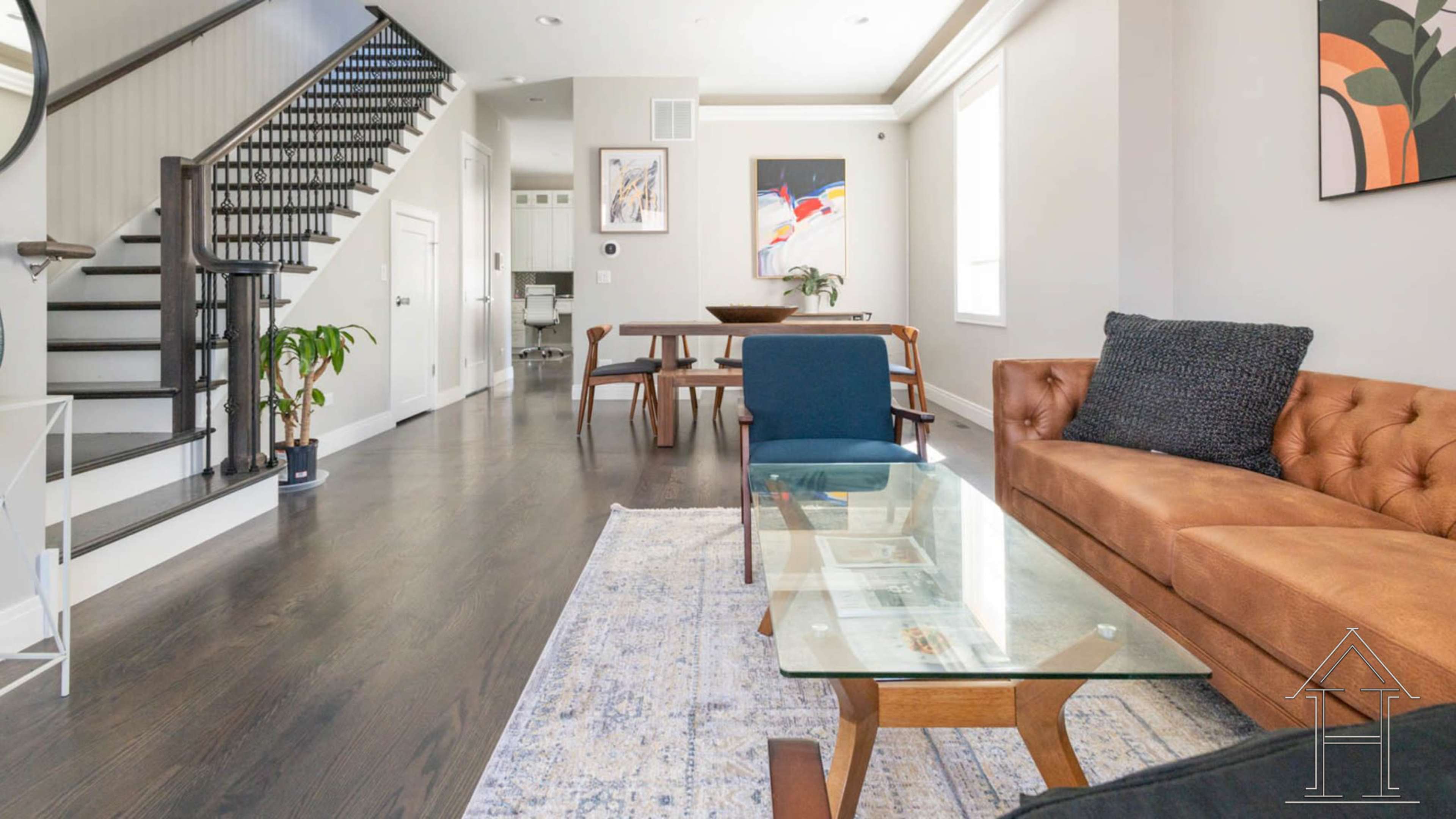 A modern living room with a brown leather sofa, a glass coffee table, and a dining area visible in the background, along with a staircase leading to an upper floor.