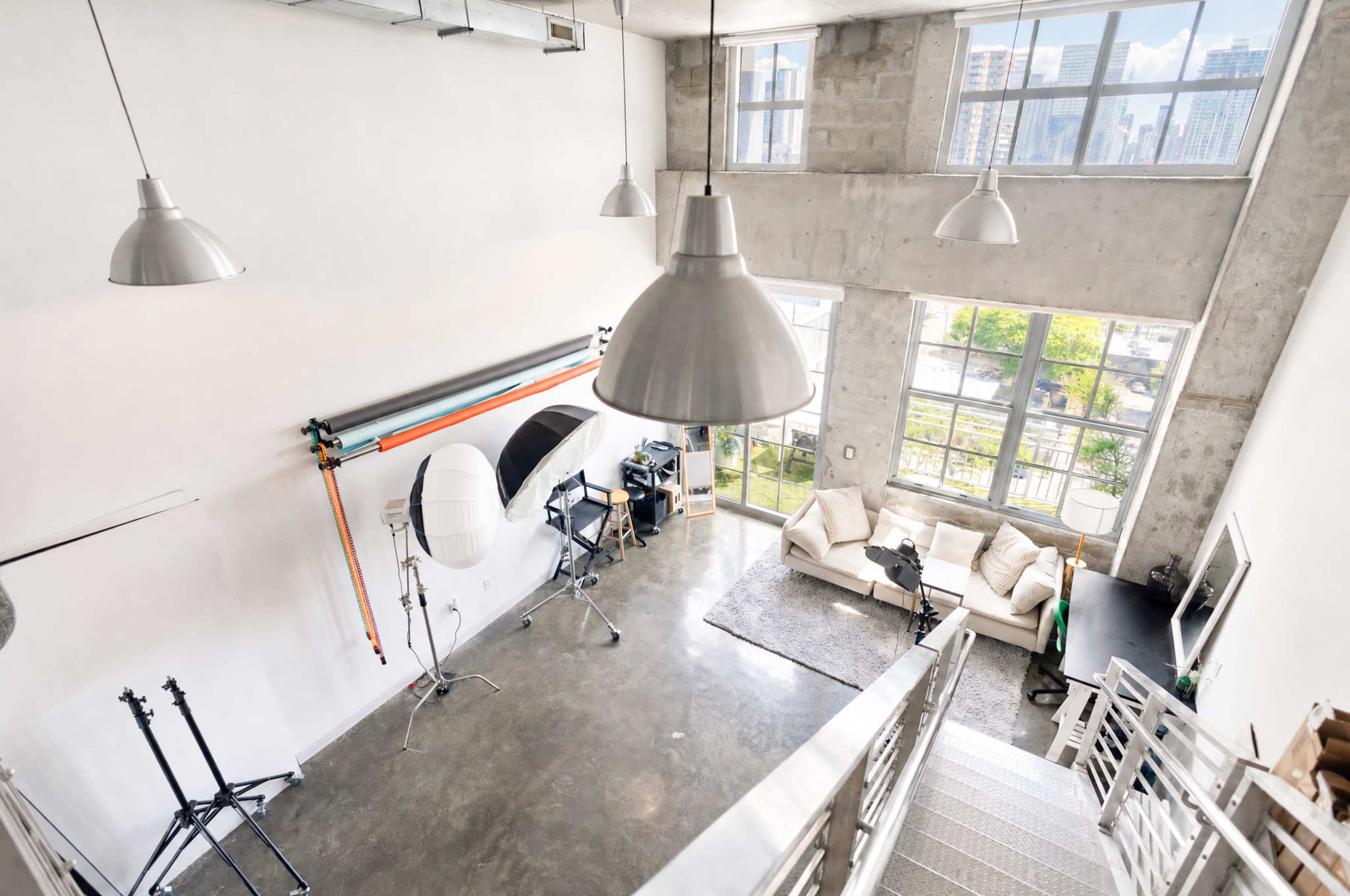 The image shows a spacious, modern studio with high ceilings, large windows, and a seating area featuring a white sofa.
