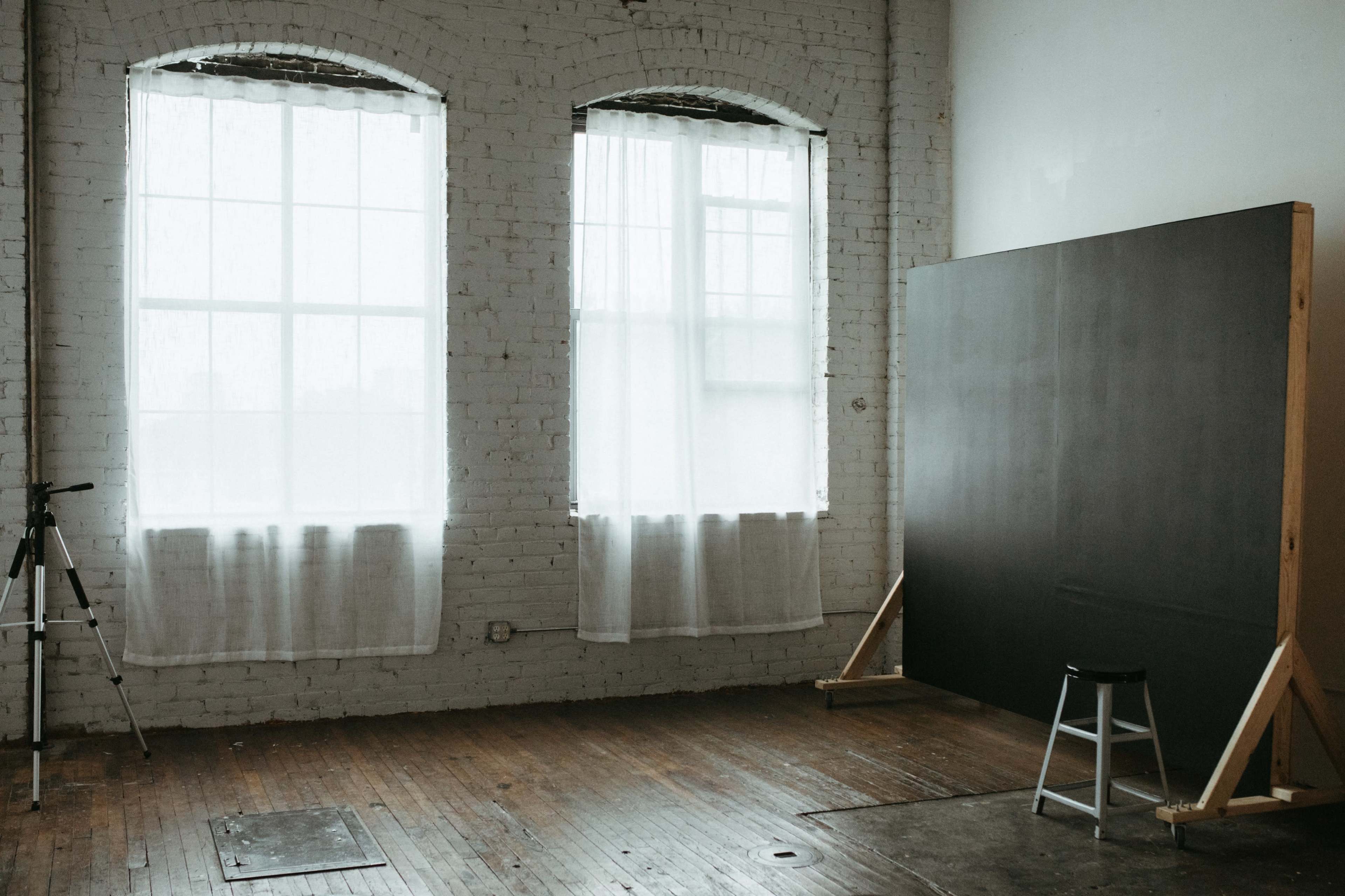 The image shows a minimalistic room with large windows draped in sheer white curtains, a large dark panel leaning against the wall, and a tripod standing nearby on the wooden floor.