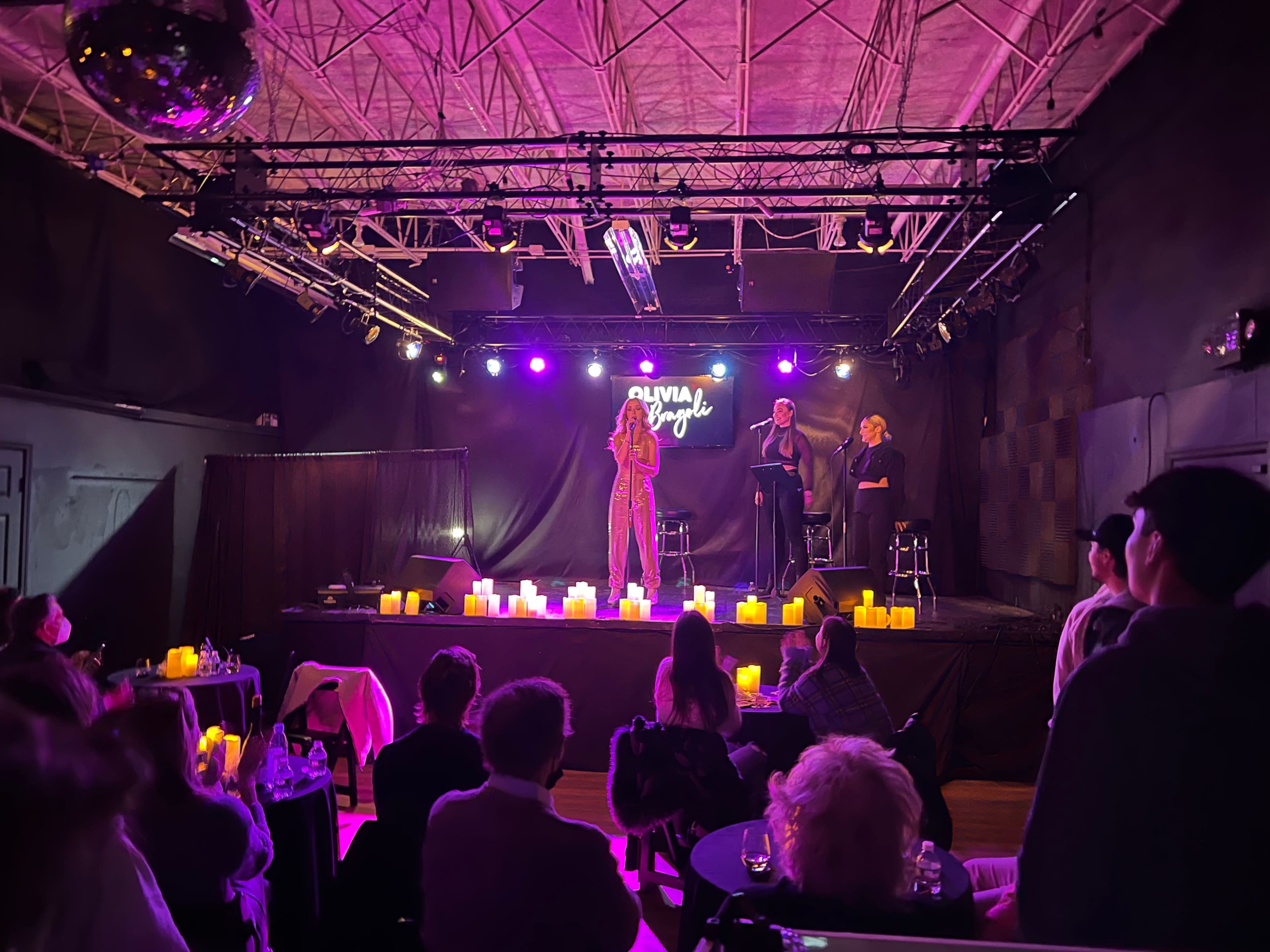 A performer sings on stage in front of an audience seated with candles lit on the floor.