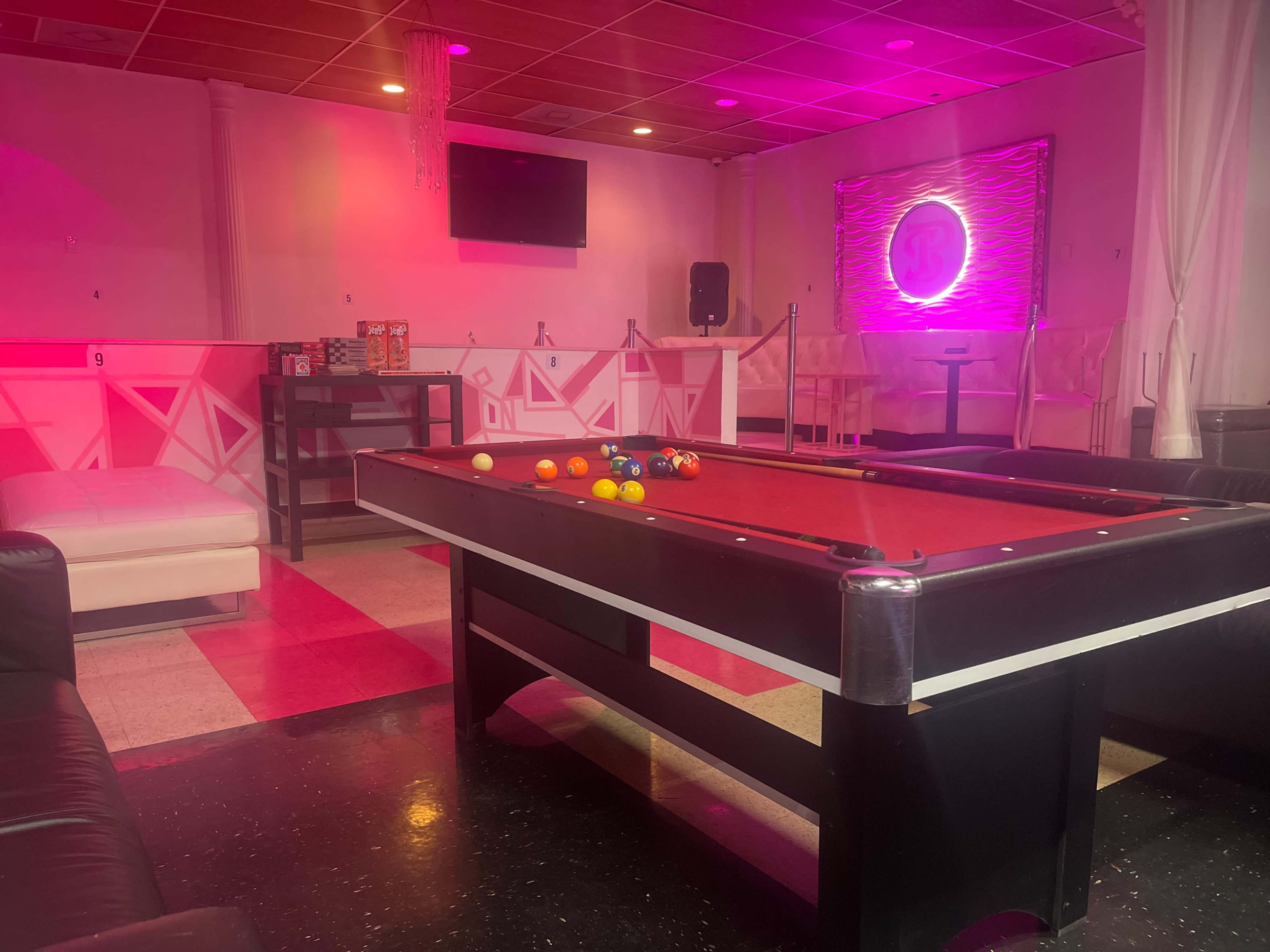 The image depicts a pool table with colorful balls arranged on it in a room featuring modern furniture and pink lighting.
