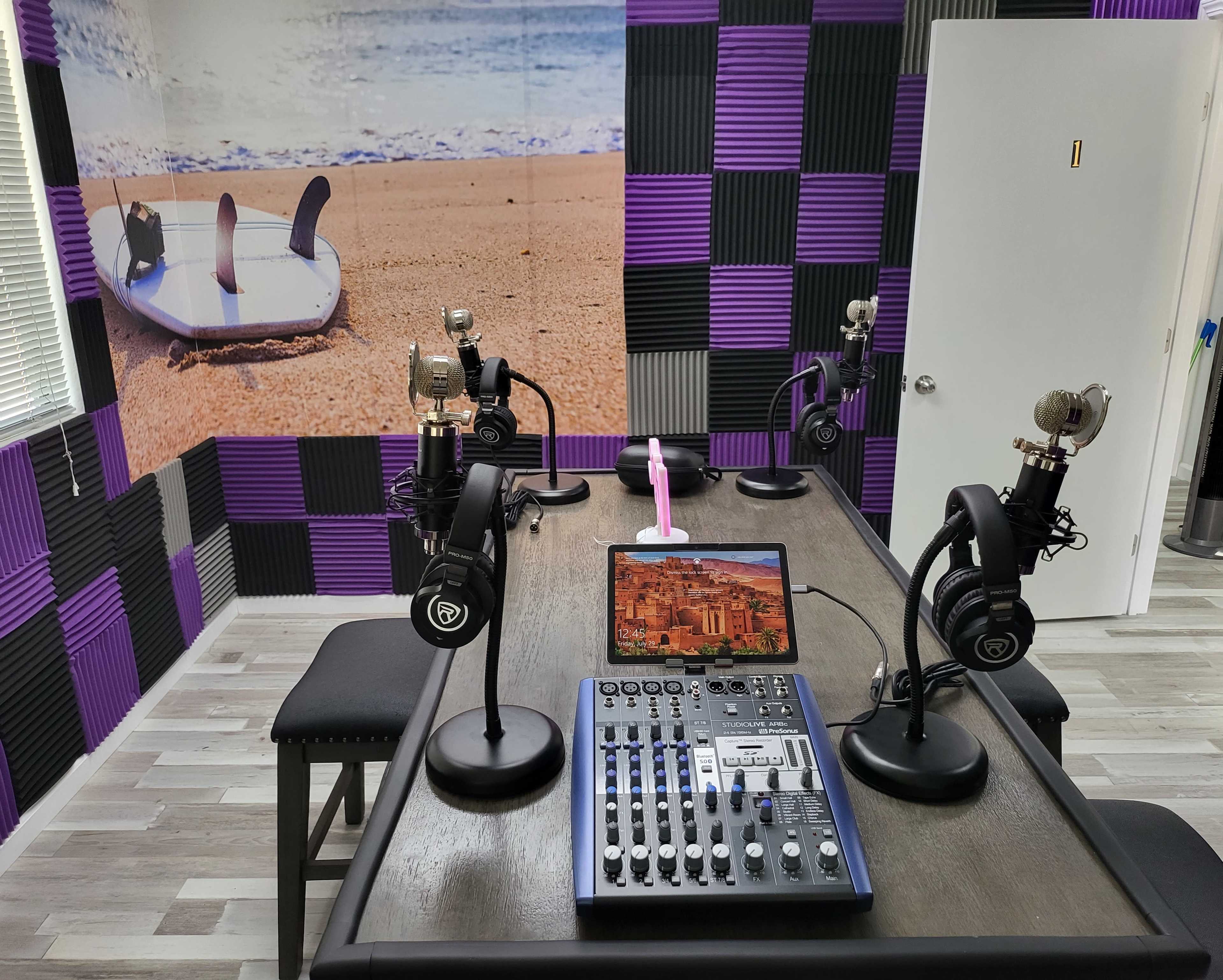 The image shows a recording studio with four microphones and a sound mixer on a table, surrounded by purple and black soundproofing panels, with a beach scene mural on the wall.