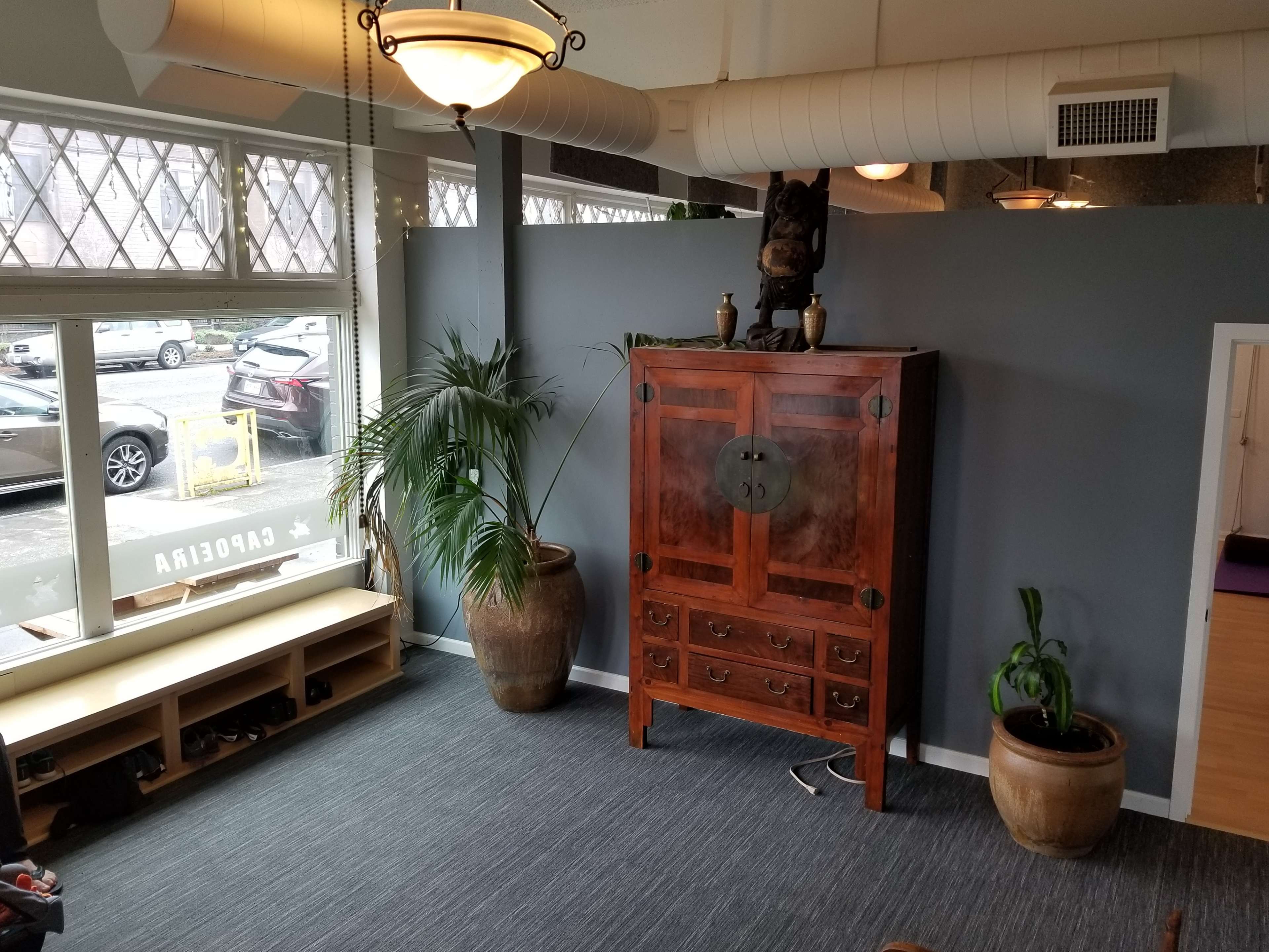 A spacious interior with a wooden cabinet against a gray wall, flanked by potted plants and a shoe rack, featuring large windows that overlook a street.