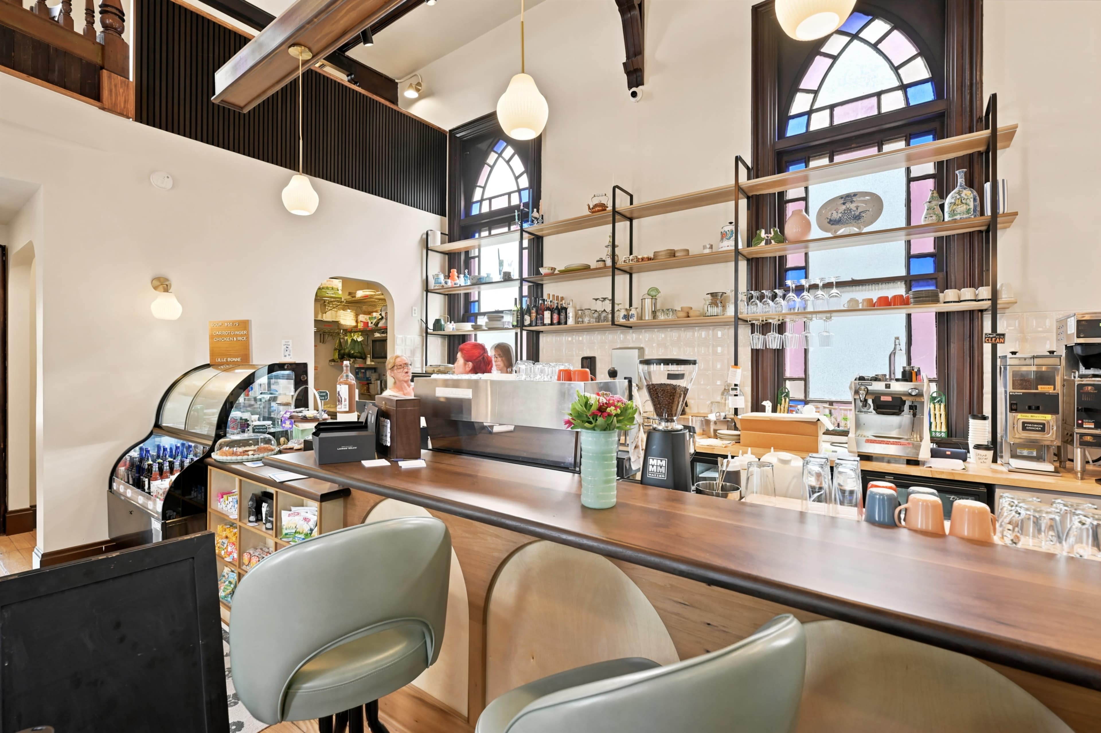 The image shows a stylish café interior featuring a wooden counter, green upholstered seating, and shelves displaying various food and drink items.