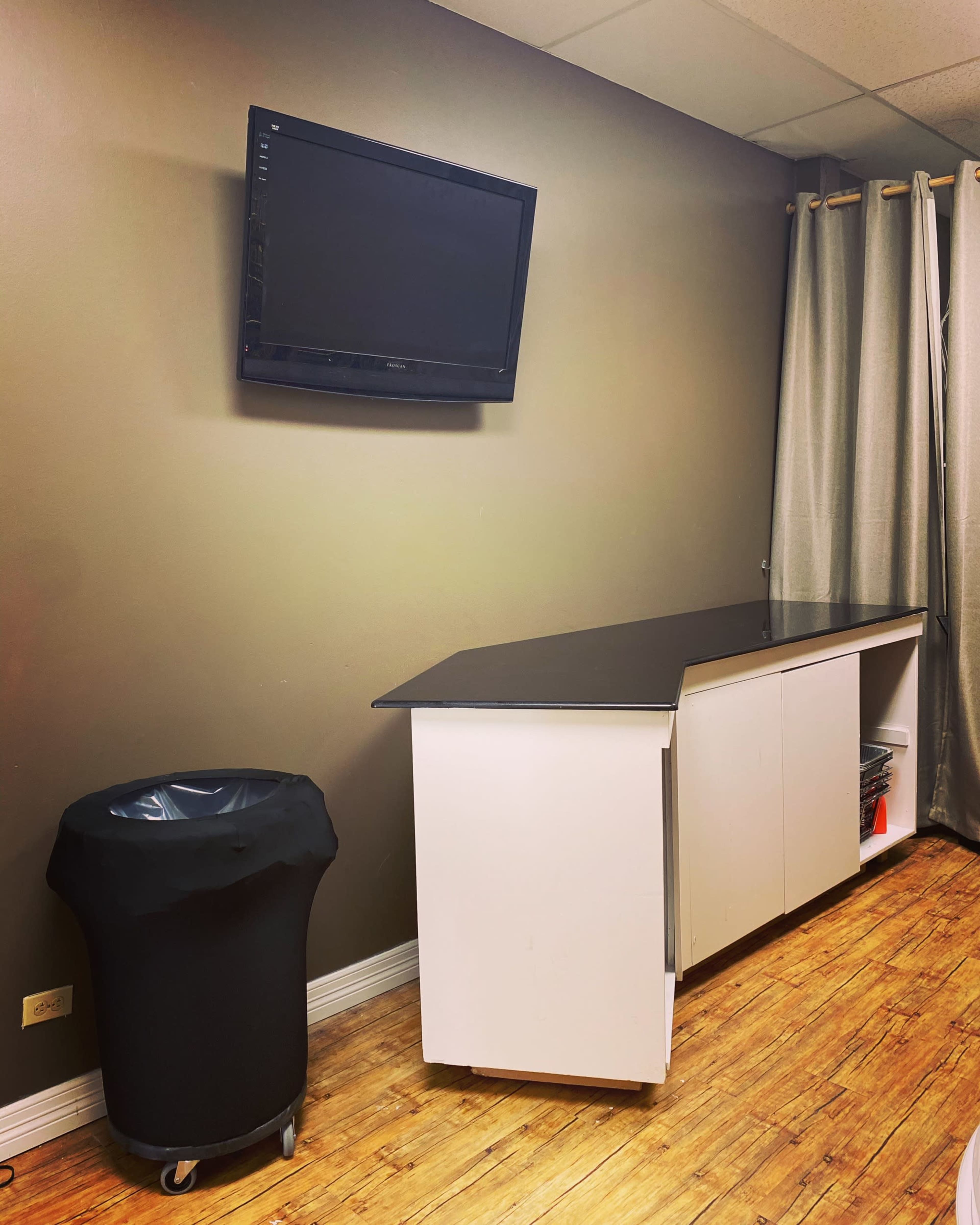 The image shows a room with a wall-mounted television, a dark trash can, and a white cabinet with a black countertop against a brown wall.