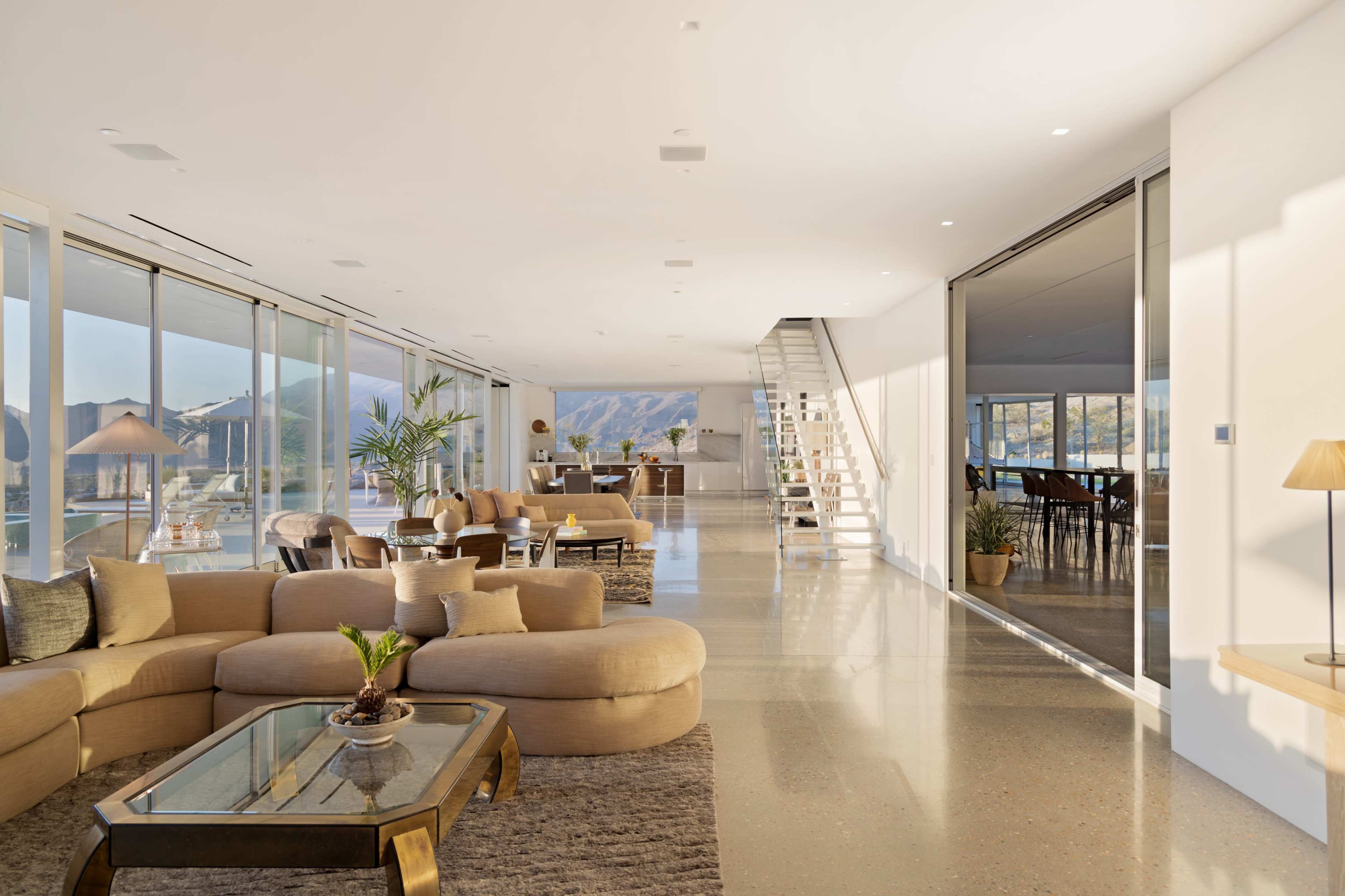 The interior of a modern, open-concept living space features a spacious lounge area with a large sofa, a glass coffee table, and large windows that offer a view of the surrounding landscape.