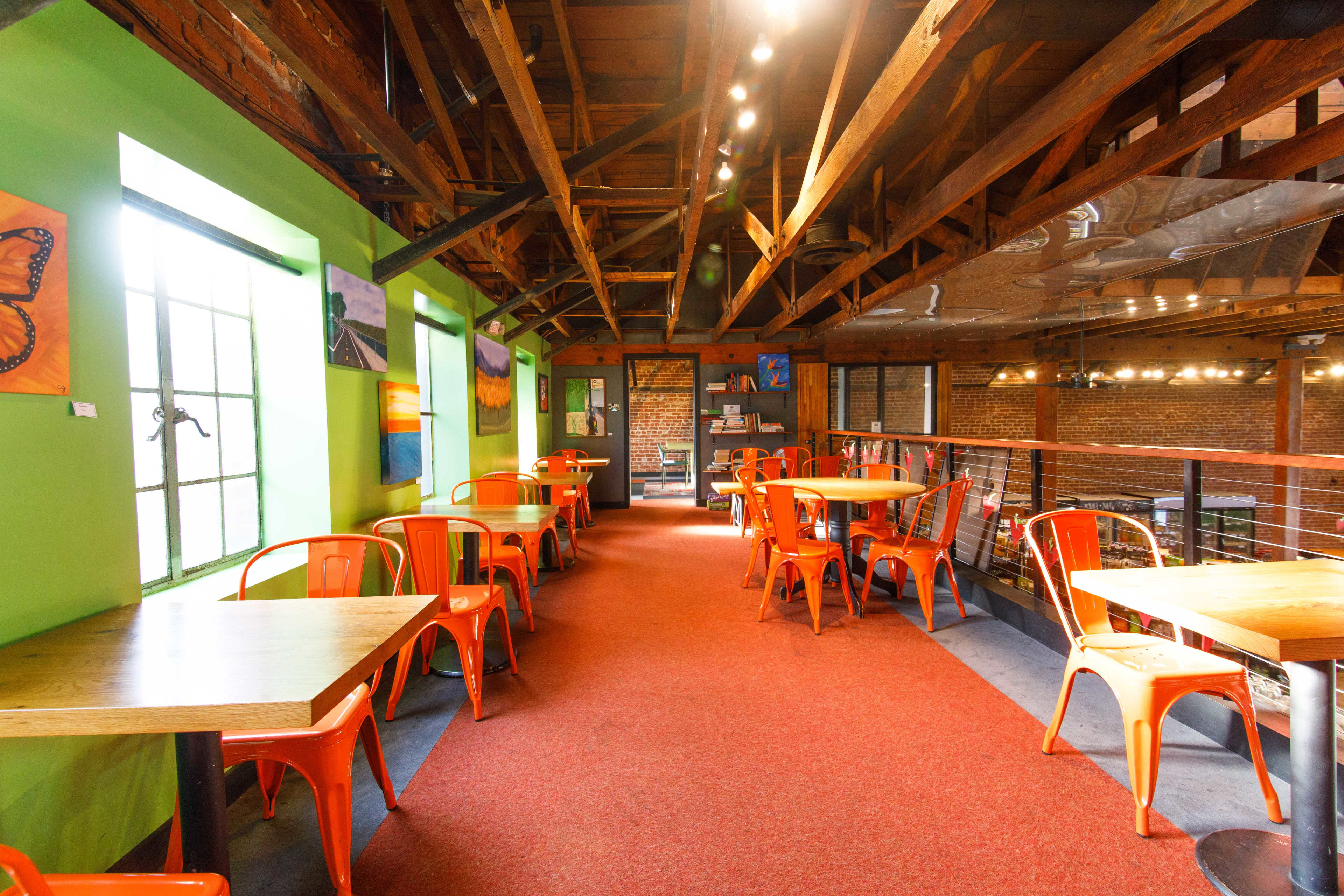 The image shows a brightly colored café with wooden beams, red carpet, and orange metal chairs arranged around wooden tables.