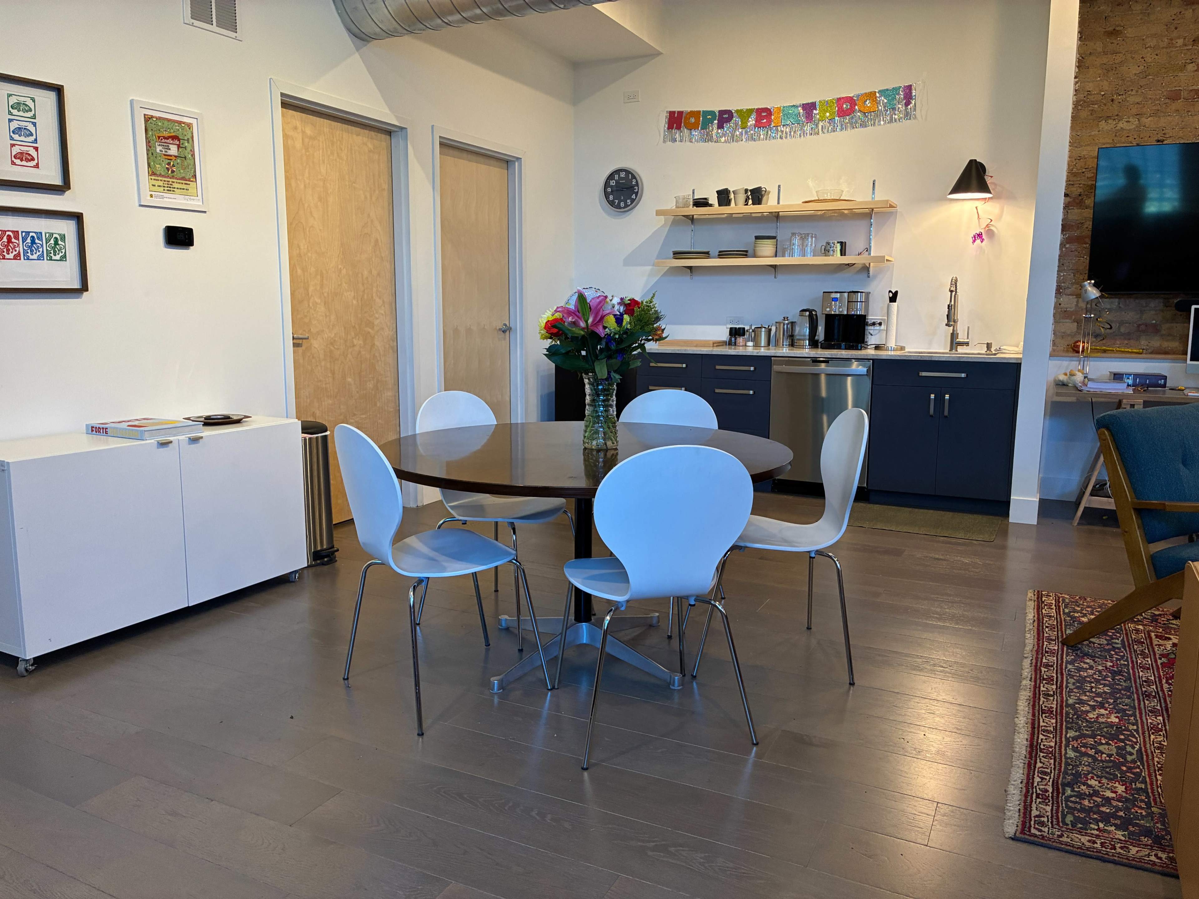 The image shows a modern dining area with a round table surrounded by four white chairs, a kitchenette in the background, and a bouquet of flowers on the table.