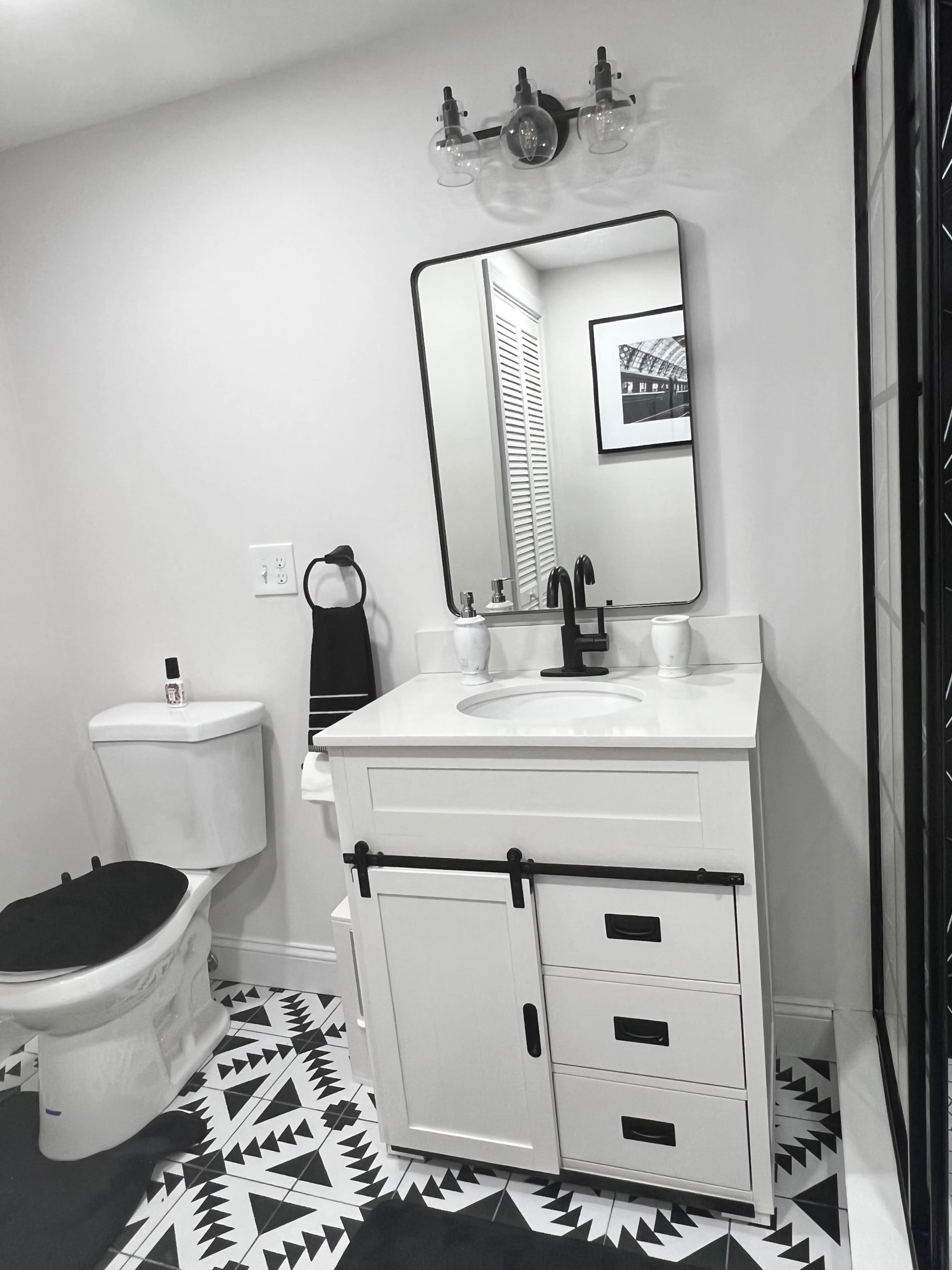A modern bathroom featuring a white vanity with a sink, a mirror, a black faucet, a toilet, and patterned black and white floor tiles.
