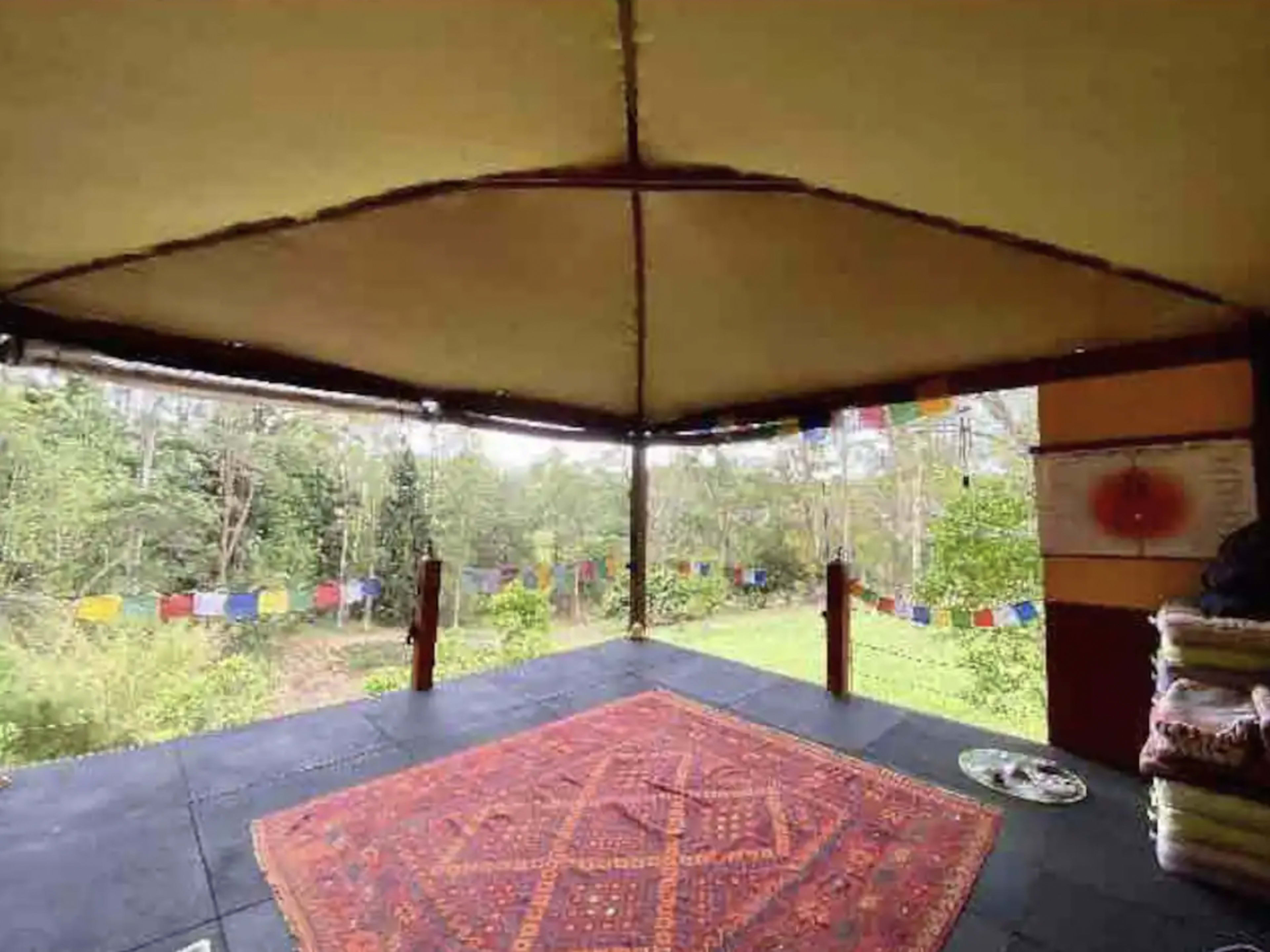 A spacious outdoor pavilion with a patterned rug and colorful prayer flags visible against a green backdrop.