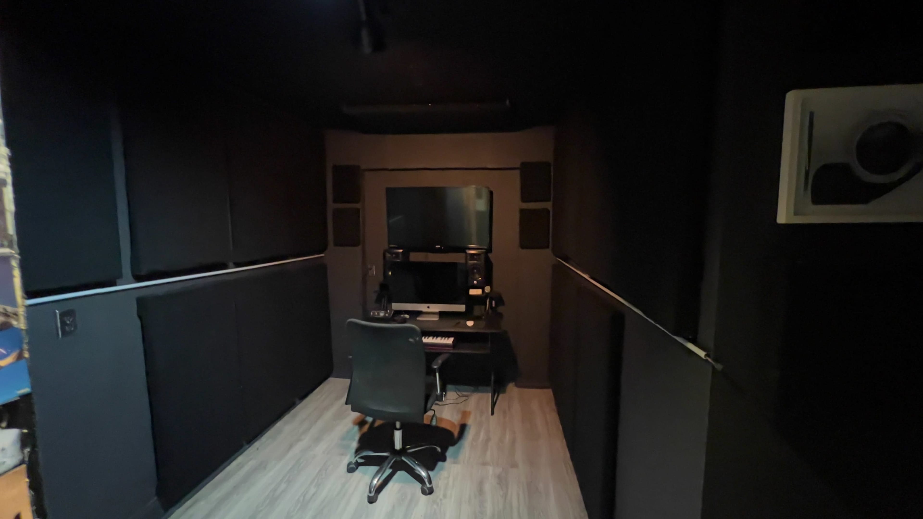 A small music studio with black soundproof walls, a computer desk, and a chair facing a monitor.
