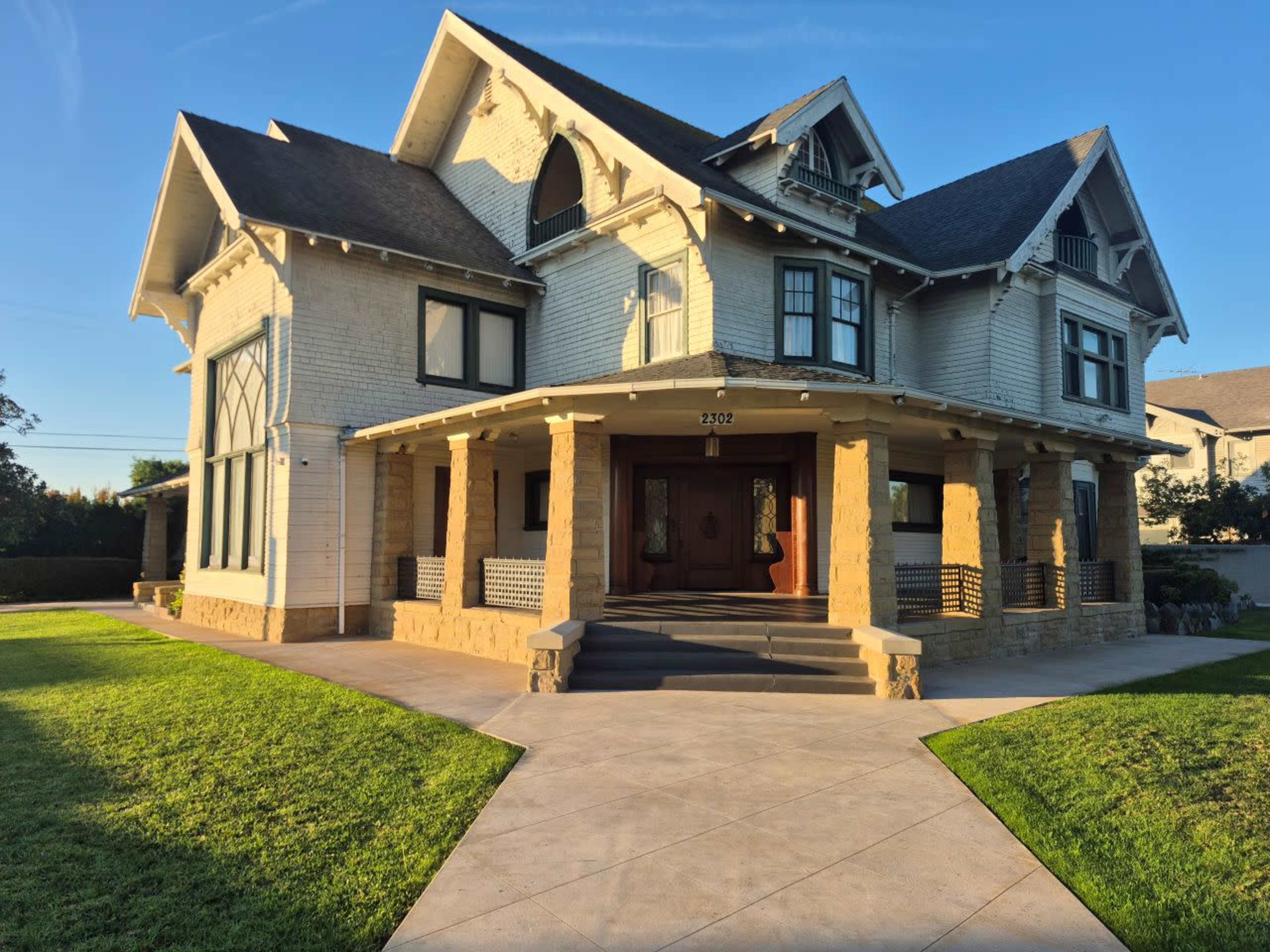The image shows a large, two-story mansion with a white exterior, decorative elements, and a front porch supported by stone columns, set in a well-maintained yard.