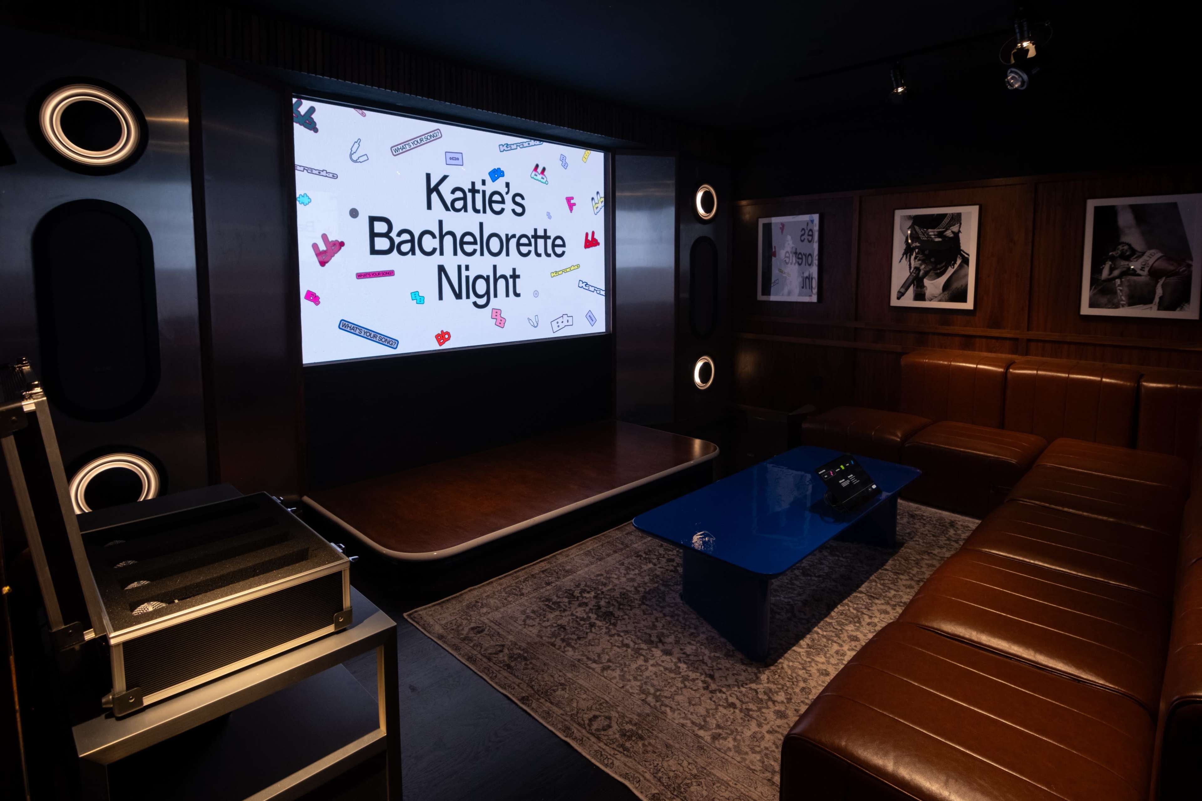 The scene shows a cozy lounge with a large screen displaying "Katie's Bachelorette Night," surrounded by brown leather seating and a blue coffee table.