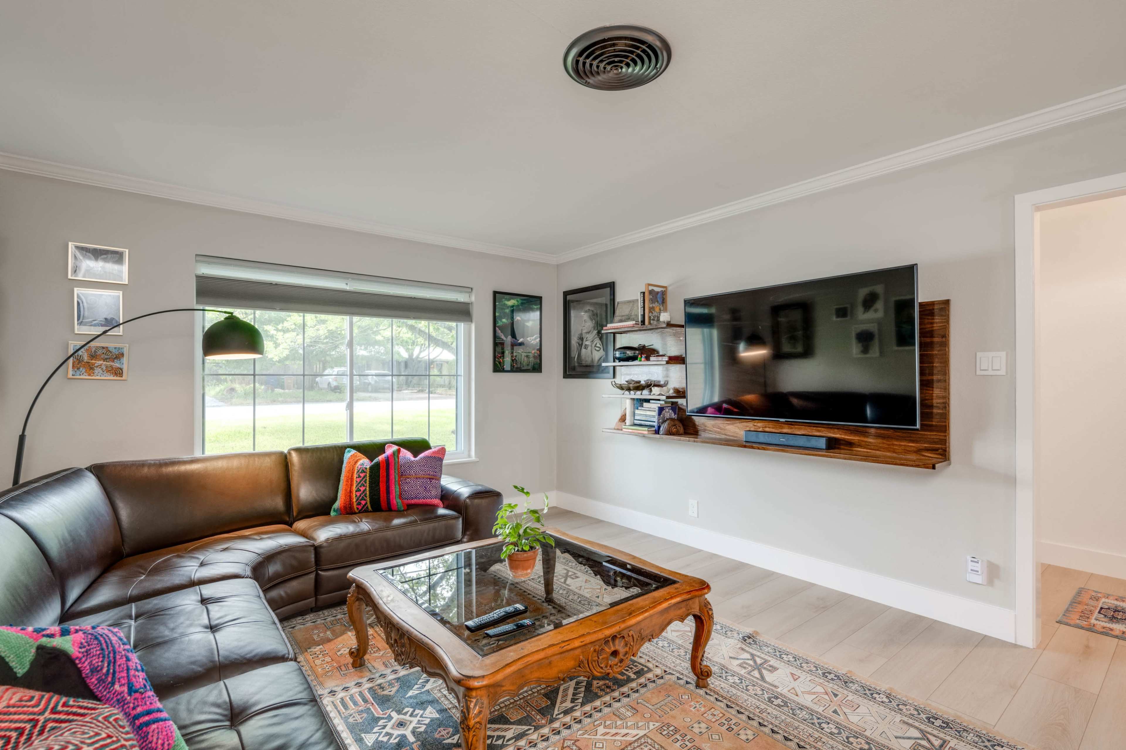 The living room features a leather sectional couch, a wooden coffee table, and a mounted television, with large windows providing natural light.