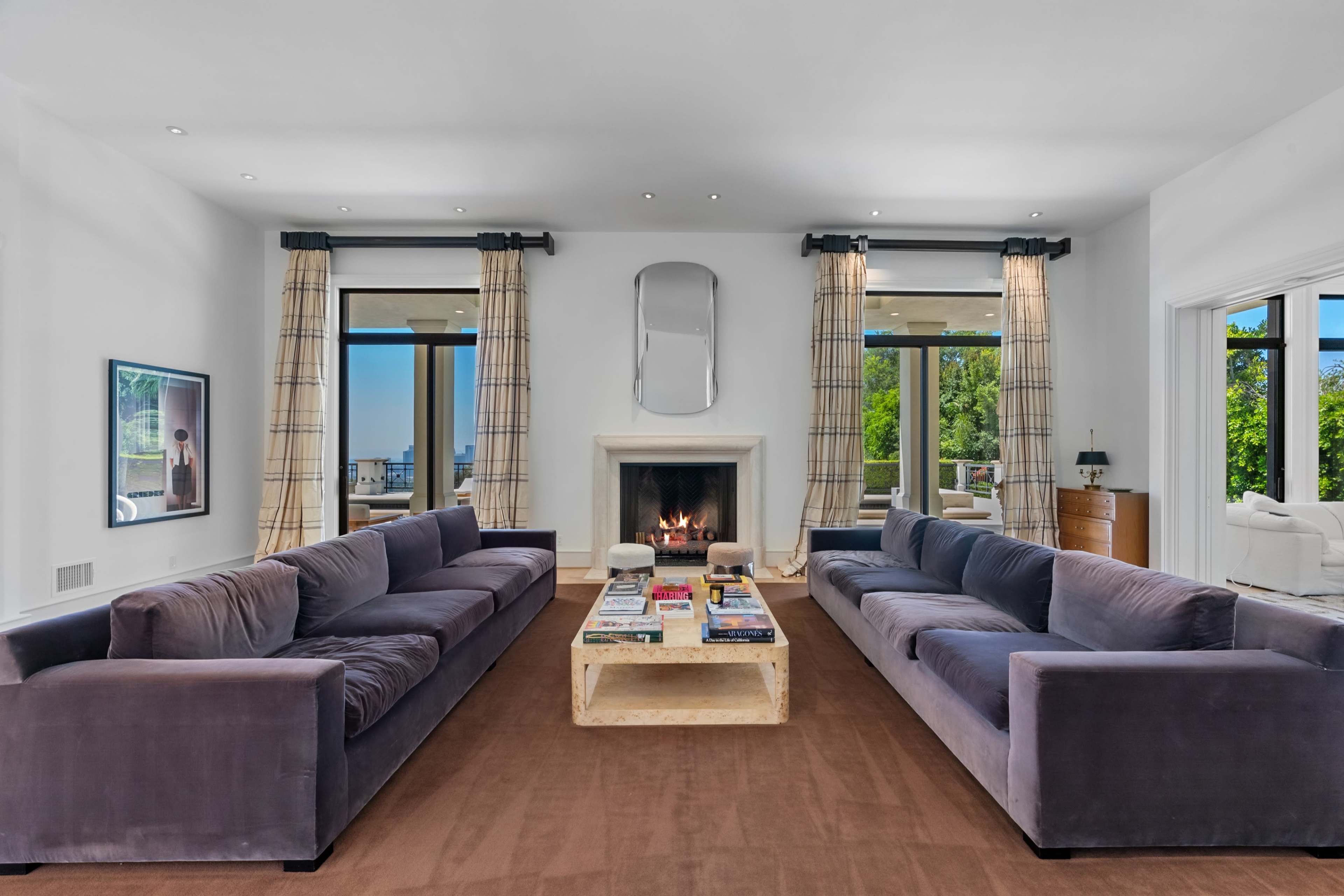 A modern living room with two large sofas facing a fireplace, framed by large windows and light-colored curtains.