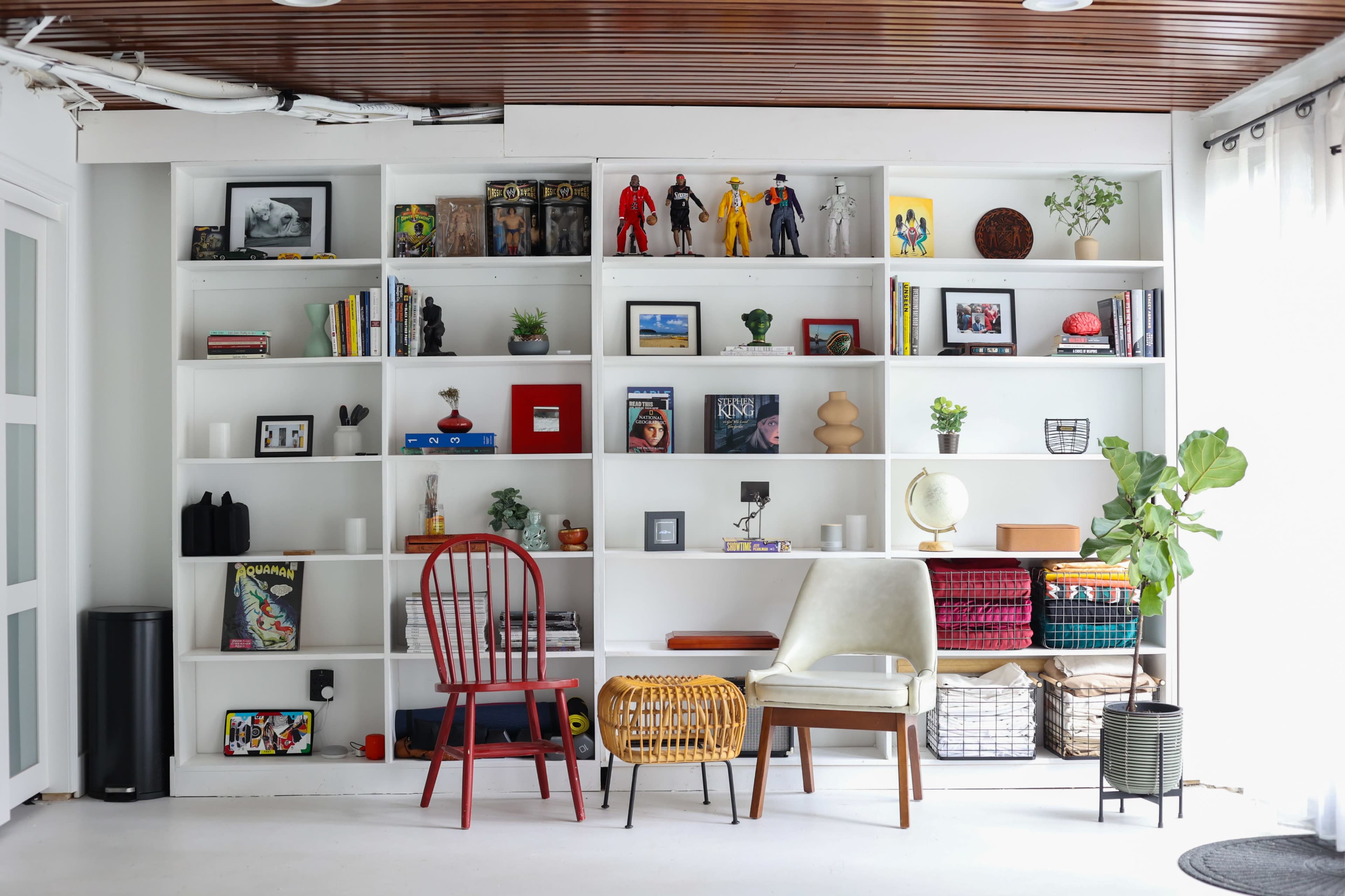 The image shows a neatly organized shelf displaying books, decorative items, and various figurines, accompanied by chairs and plants in a bright room.
