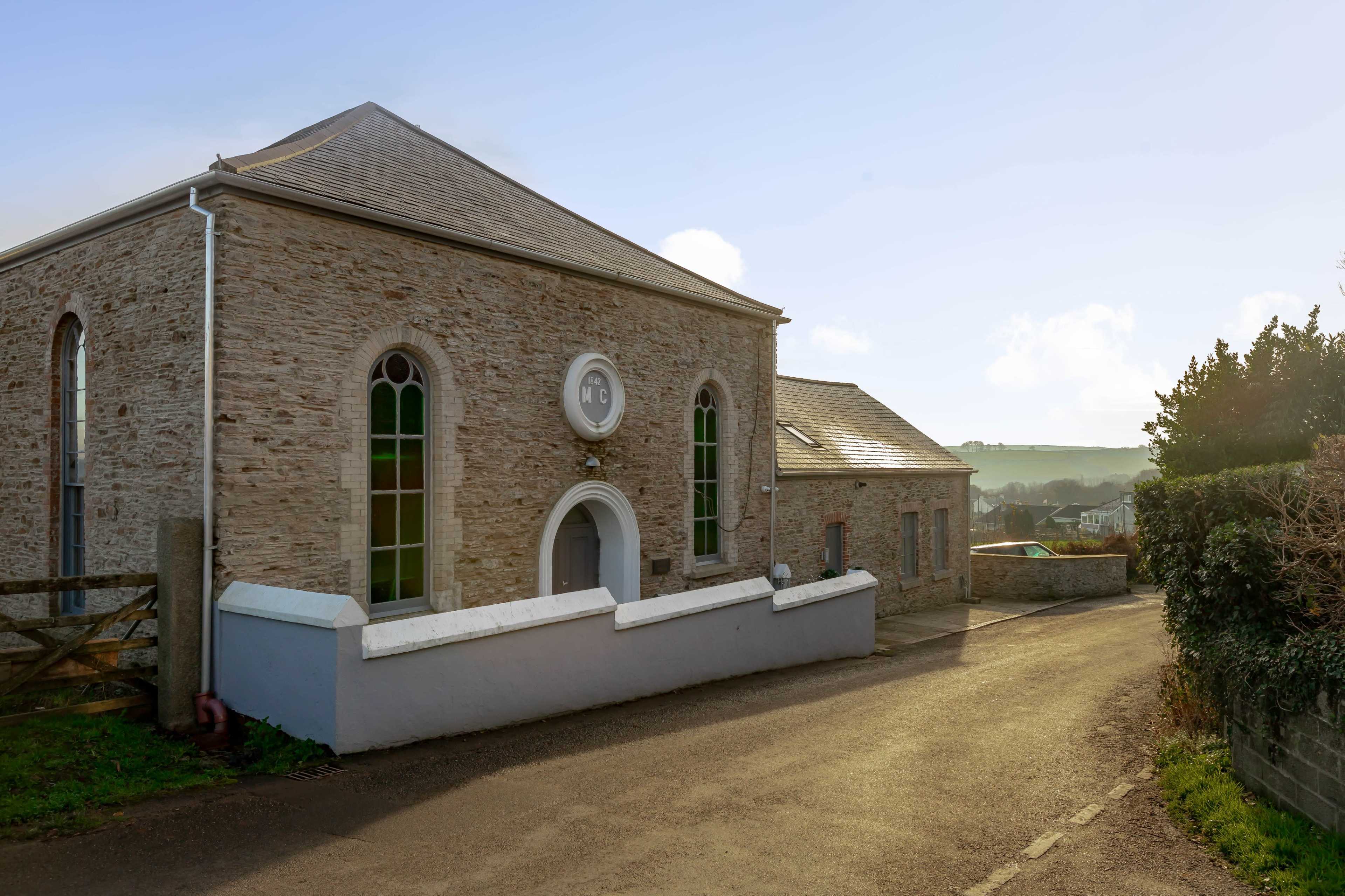 A stone building with arched windows and a sloped roof, situated beside a gravel road surrounded by greenery.