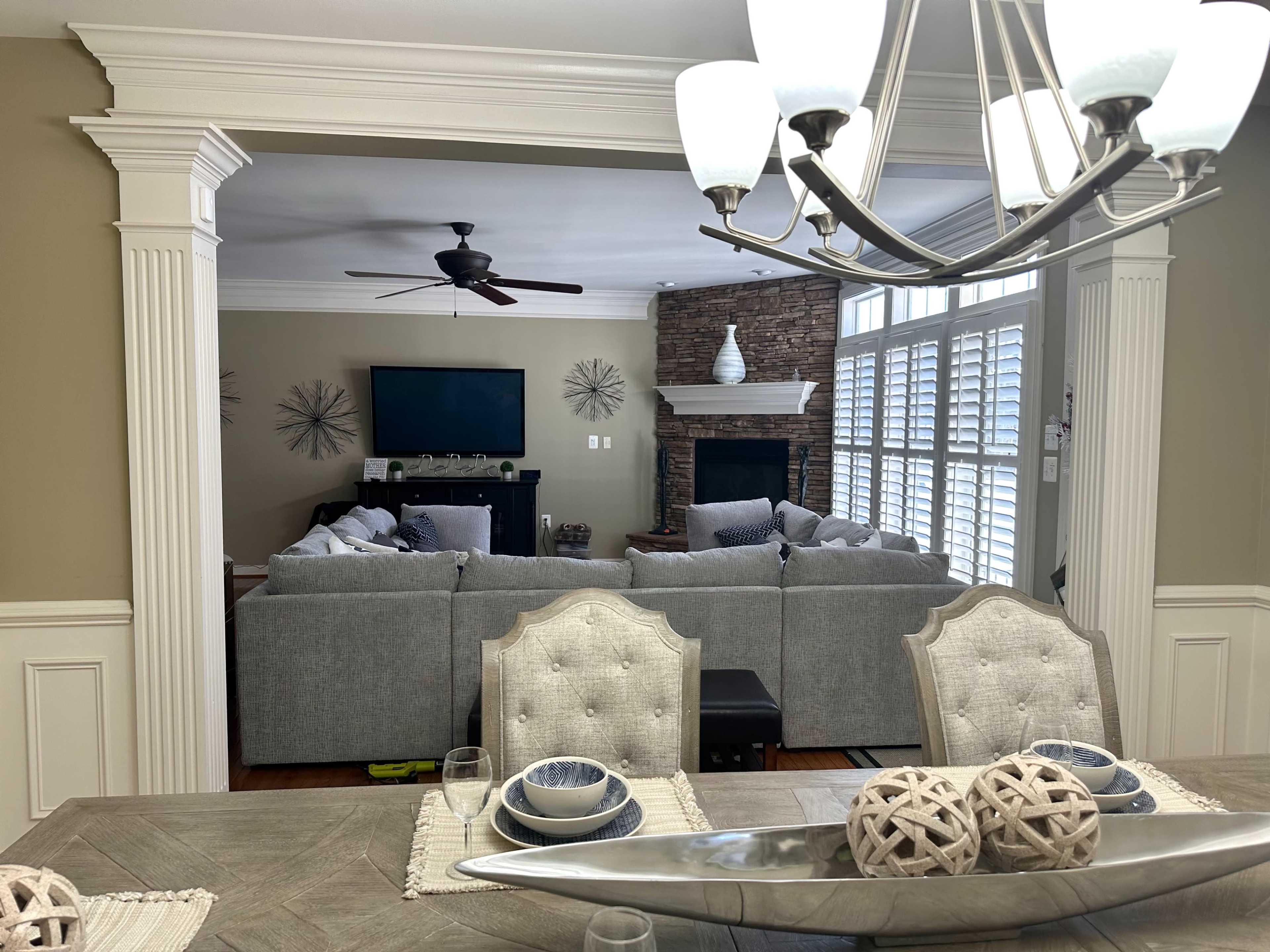 The image shows a dining area with a table set for meals, looking into a living room featuring a stone fireplace and large gray sofas.